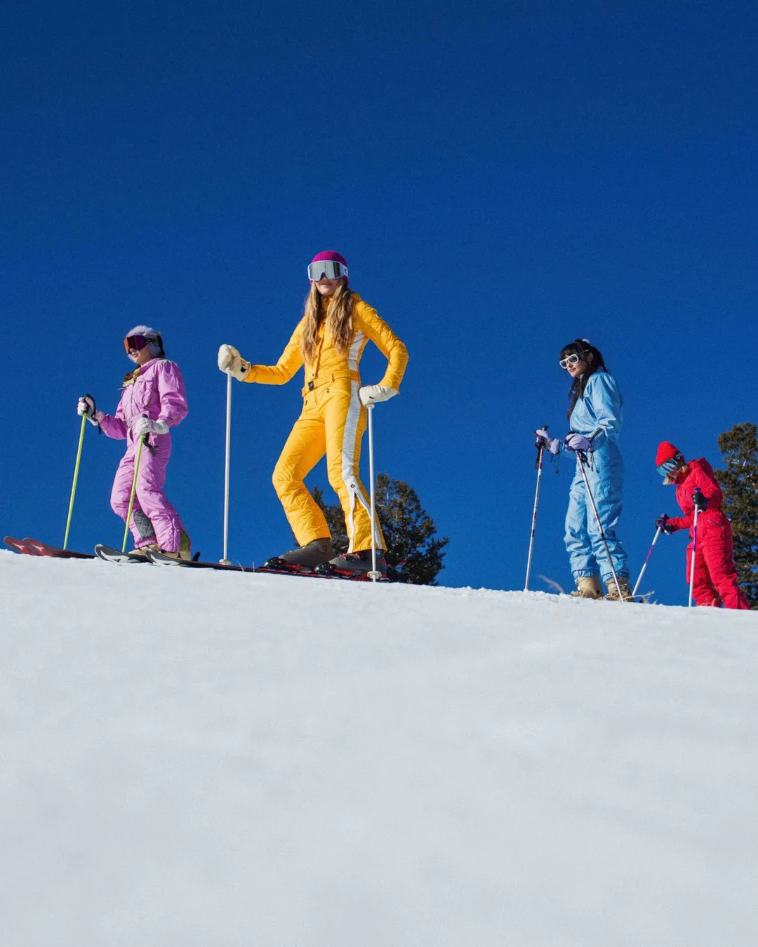 Group of five children skiing on snow-covered slope, wearing colorful ski suits and goggles, with clear blue sky and trees in the background.