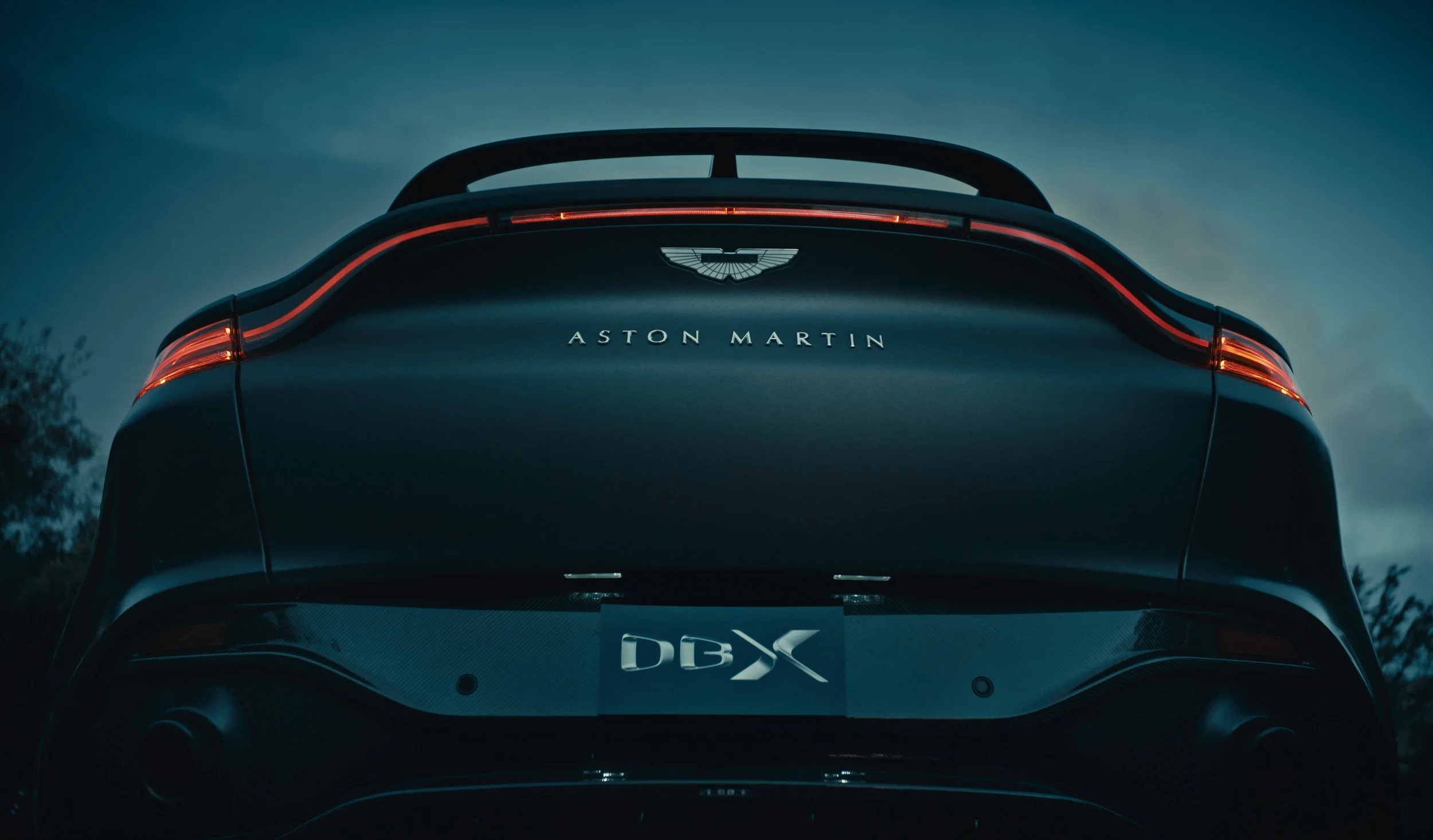 Rear view of a dark blue Aston Martin DBX, with illuminated tail lights and the Aston Martin logo on the trunk.