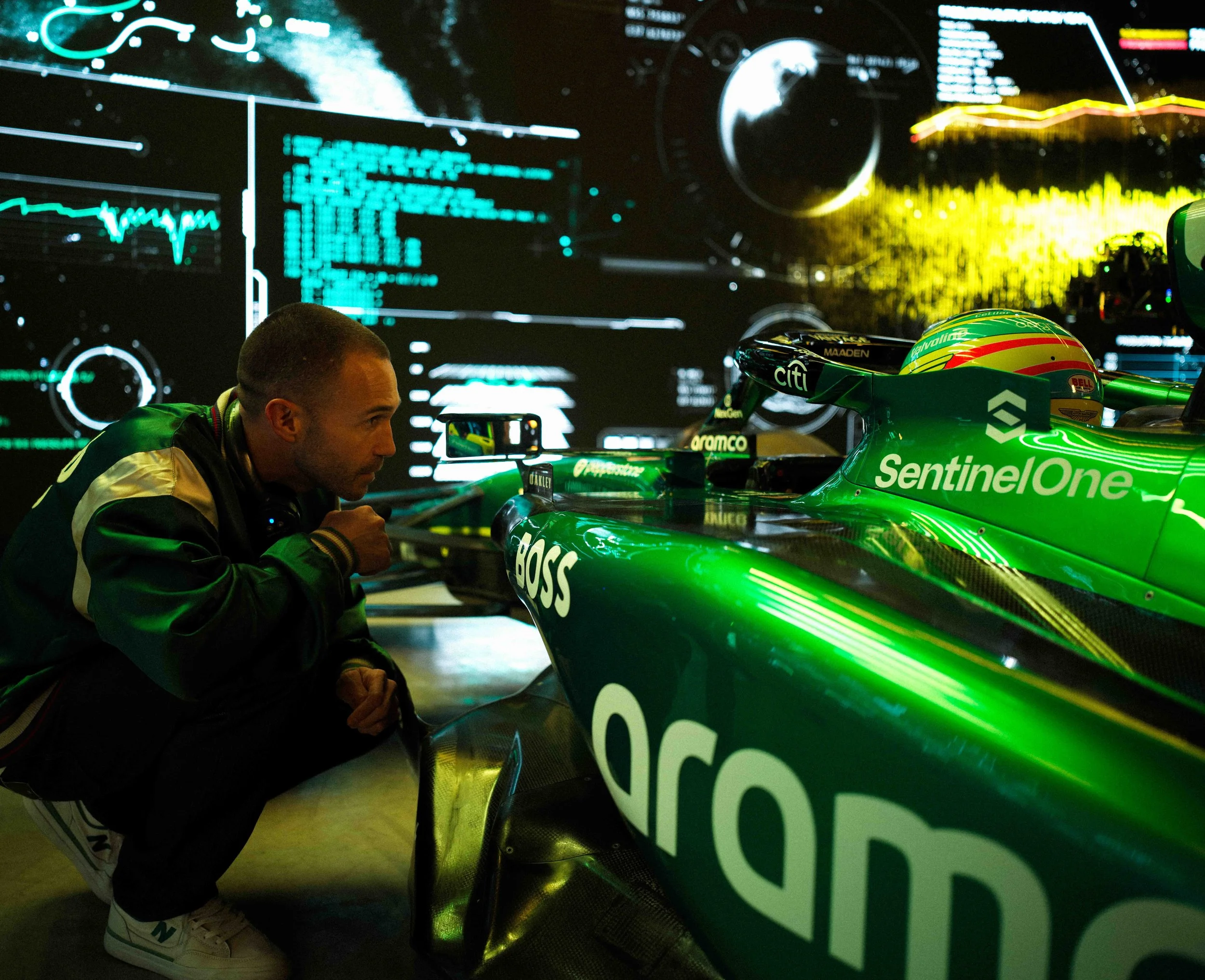 A man kneeling next to a green racing car with sponsor logos, looking at it in a dimly lit room with digital screens displaying data and graphics in the background.