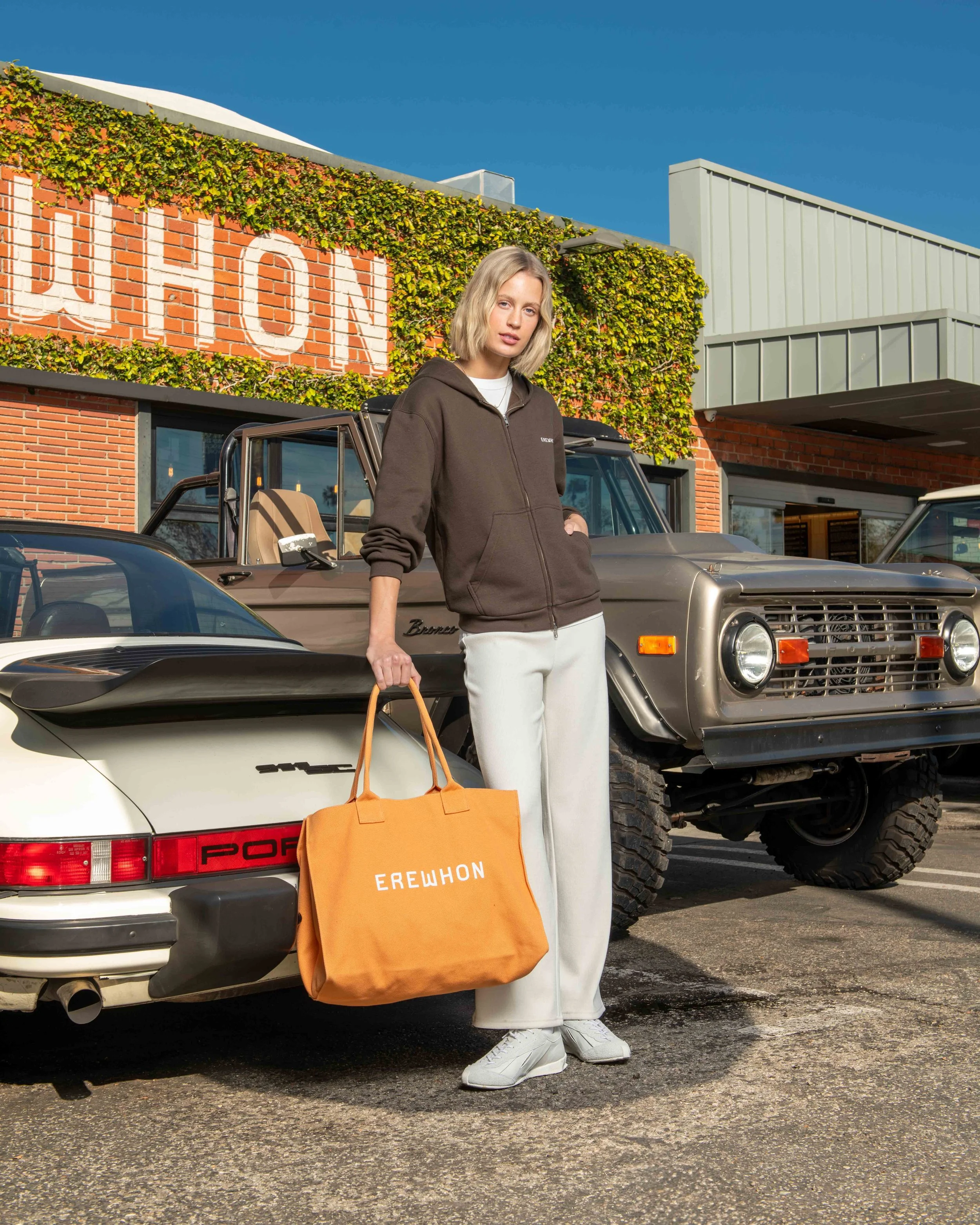 A woman standing in a parking lot between two vintage cars in front of a building with a brick and ivy exterior. She is holding an orange tote bag with white lettering and is dressed in a brown hoodie, white pants, and white sneakers.