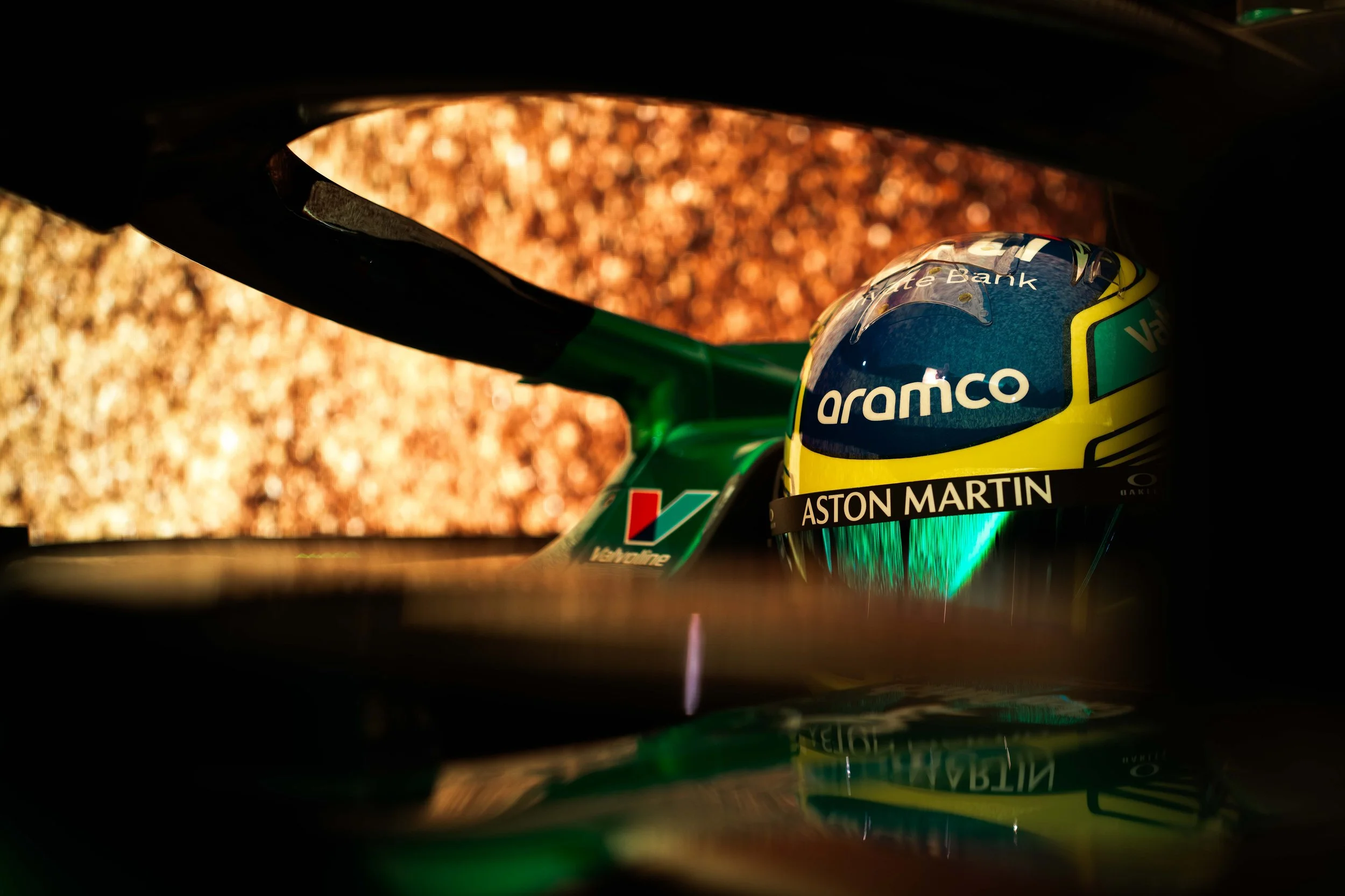 A racing helmet with Aston Martin branding, placed on a car dashboard, with a blurred background featuring a textured, gold-colored surface.