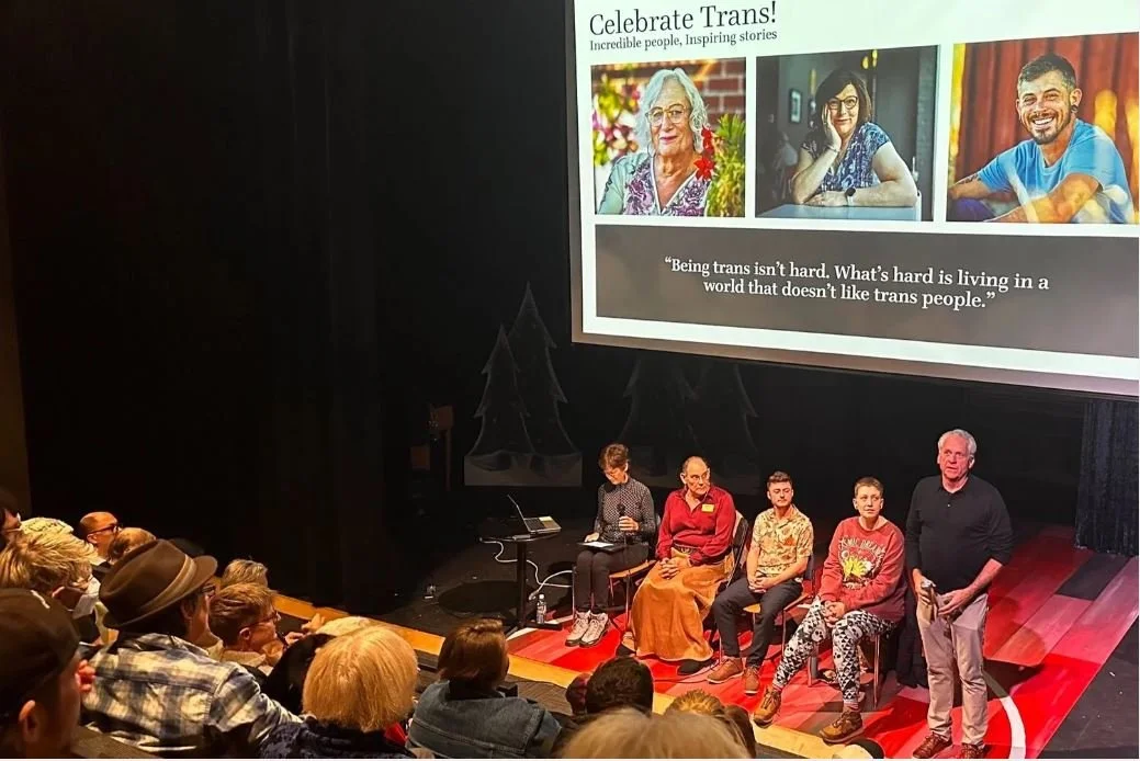 A stage containing 5 panel speakers is in the photo. The speakers stand in front of a projector displaying the words, "Being trans isn't hard. What's hard is living in a world that doesn't like trans people." From 2024 Trans Proud event in Boulder