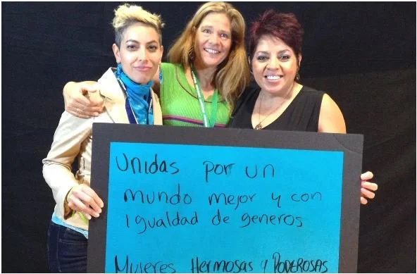 Three smiling females hold a blue sign that says "Unidas por un mundo mejor y con igualdad de generos"