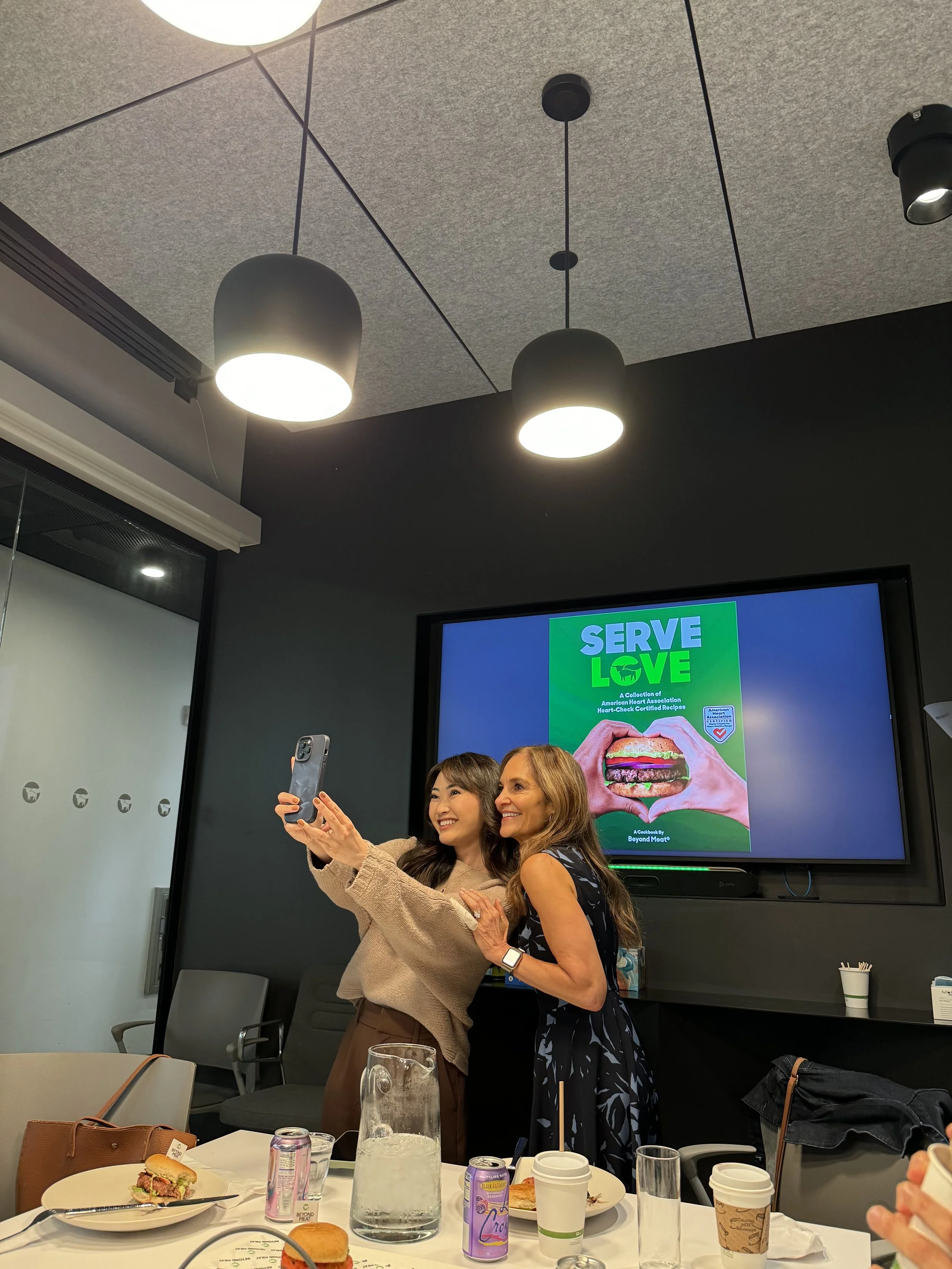 Two women taking a selfie in a conference room with a screen displaying a cookbook cover titled 'Serve Love.' The table in front has drinks, food, and utensils.