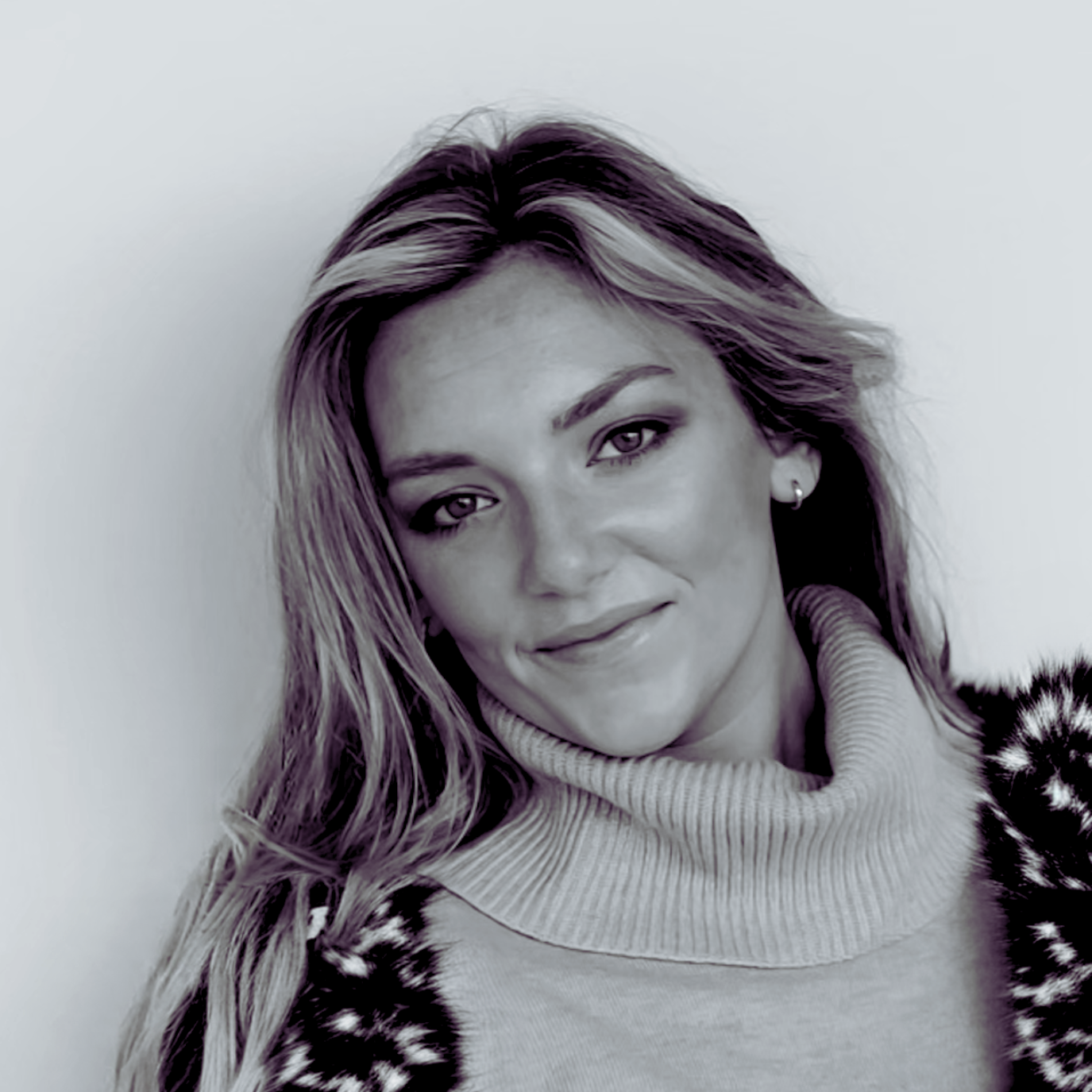 Black and white portrait of a woman with long wavy hair, wearing a turtleneck and a patterned jacket, smiling slightly and looking at the camera.