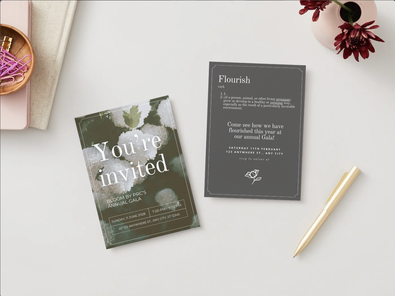 Invitation card for Bloom by PRC's annual gala with the text 'You're invited' on a background of white flowers, alongside a black program booklet, a gold pen, a pink vase with red flowers, and a pink notebook with a wooden container filled with pink paper clips on a white surface.