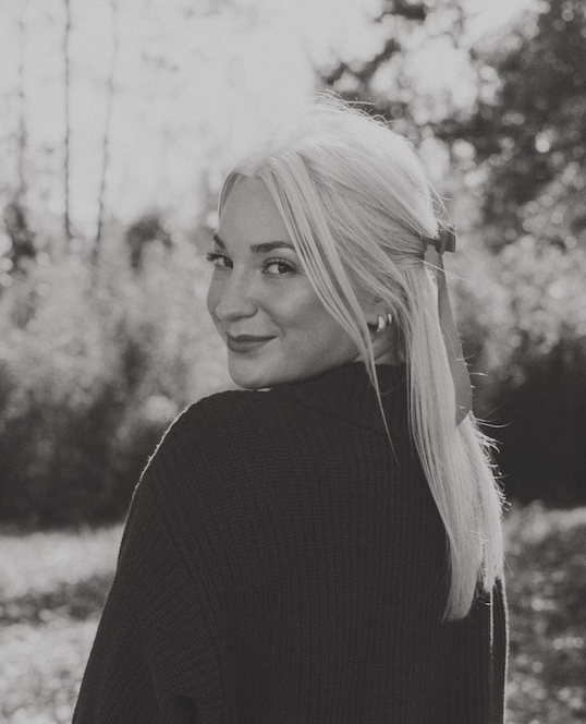 Black and white photo of a blonde woman with a hair clip, looking over her shoulder outdoors.