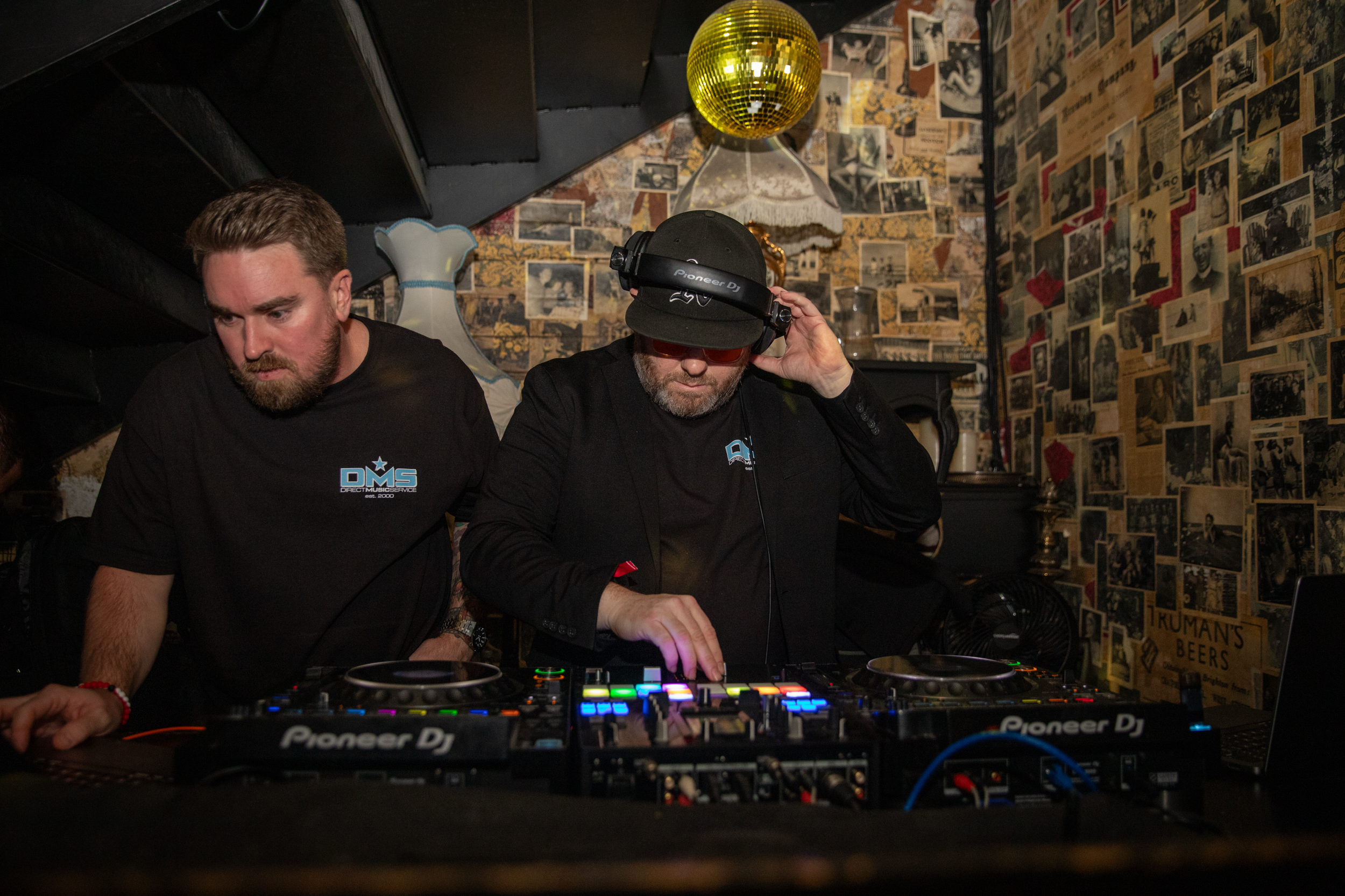 Drew and Fuseamania DJing at a nightclub in Vegas, one wearing a black jacket and headphones, the other in a black T-shirt with 'DMS' logo, surrounded by old photos and a gold disco ball