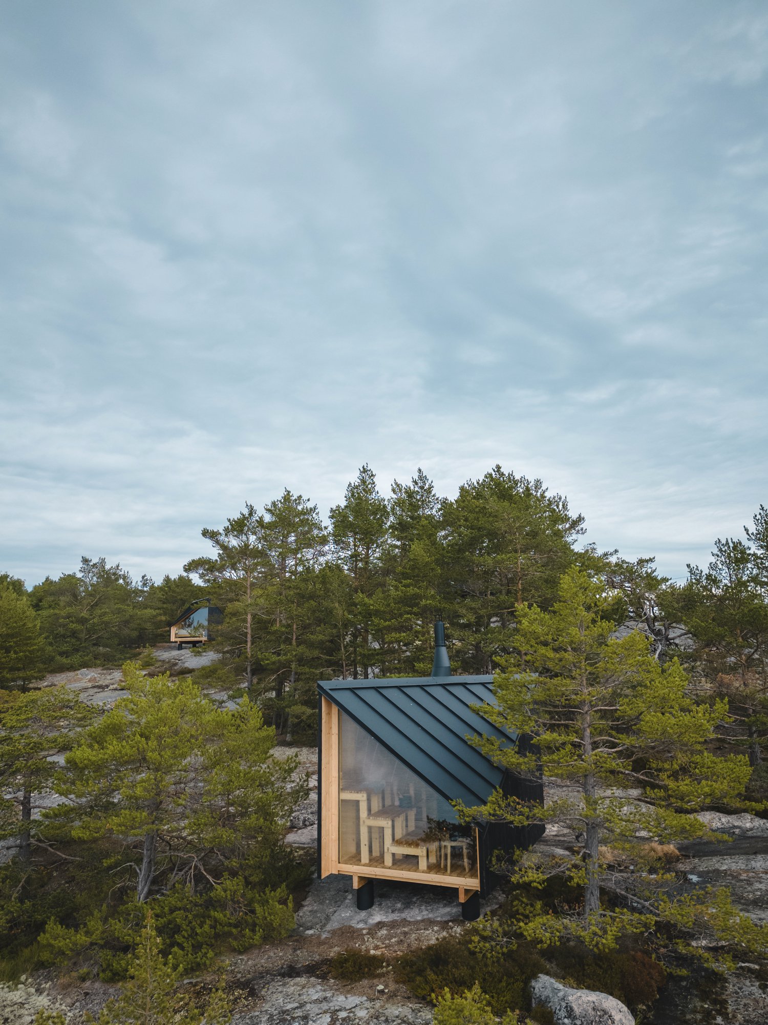 Modern cabin with slanted roof and large windows in a forested area, surrounded by trees and rocky terrain under a cloudy sky. Space of Mind Cabin, available in the USA. Real Finnish Sauna. Hotel Cabin, for Hospitality projects.