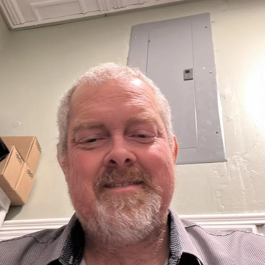 A smiling middle-aged man with gray hair and a beard taking a selfie indoors. The background shows a gray electrical panel on a light-colored wall and some cardboard boxes on a shelf.