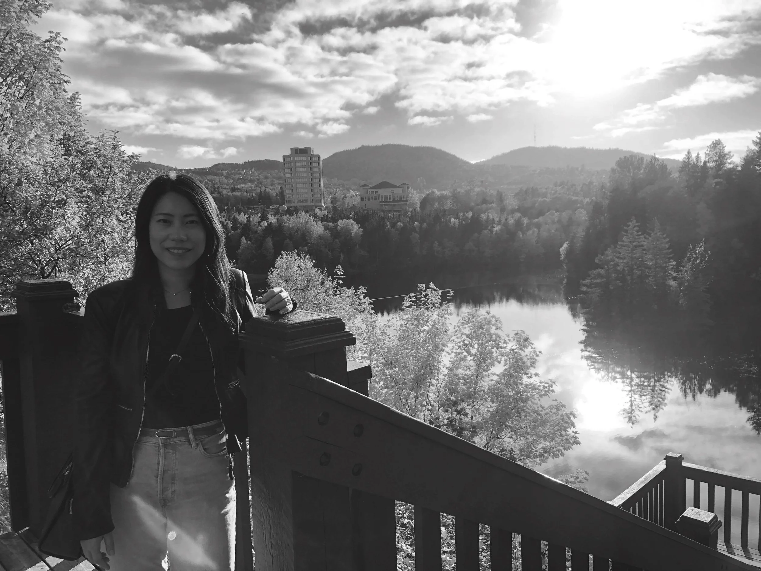 A woman standing on a bridge overlooking a river and a landscape with trees, buildings, and mountains in the distance, in black and white.