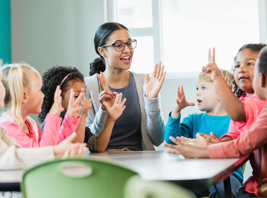 A teacher engaging with a diverse group of young children during a classroom activity. They are sitting around a table, using hand gestures, smiling, and interacting enthusiastically.