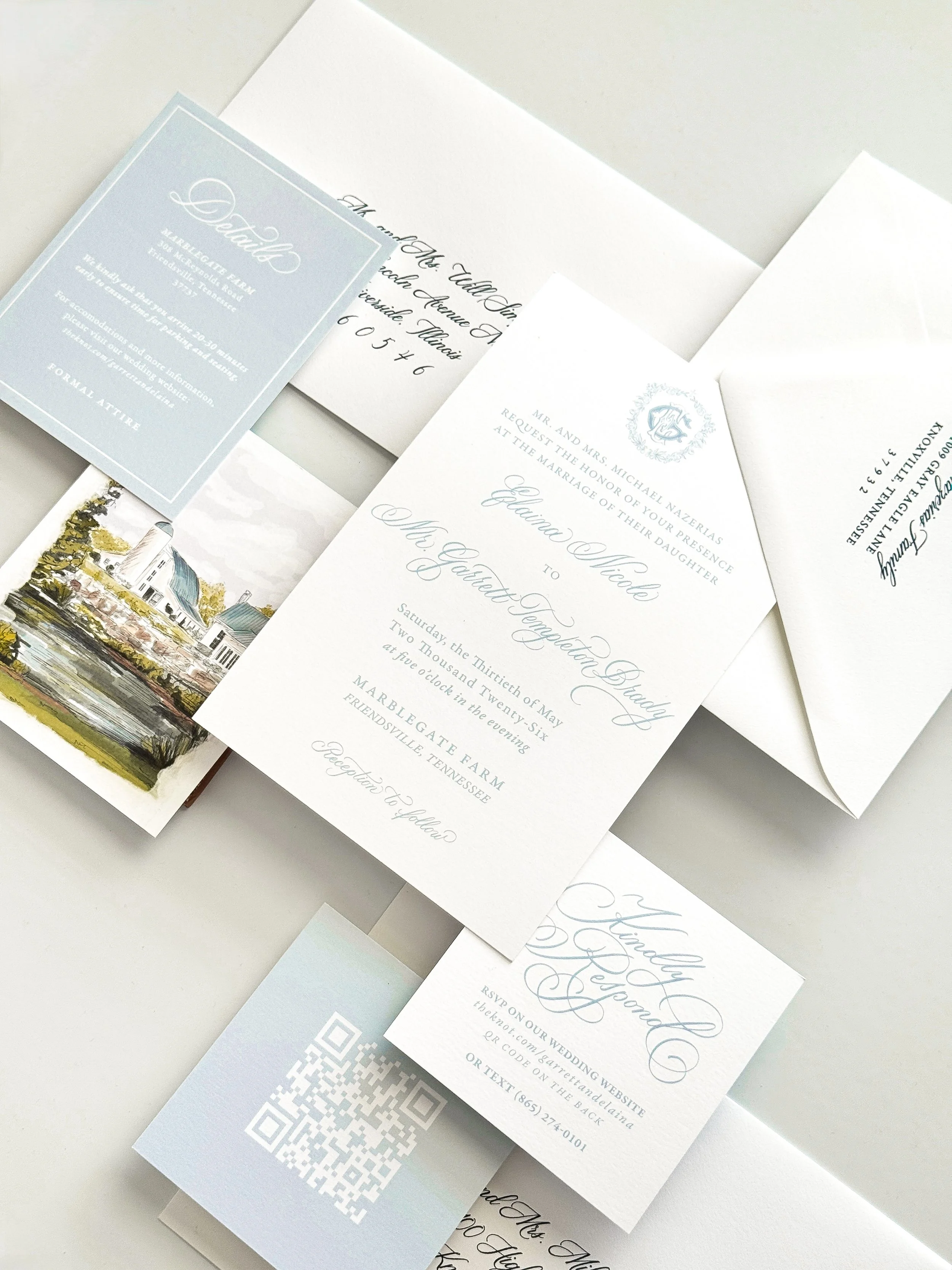 Various wedding invitations laid out, including a card with a QR code and event details, and an illustration of a house with trees.