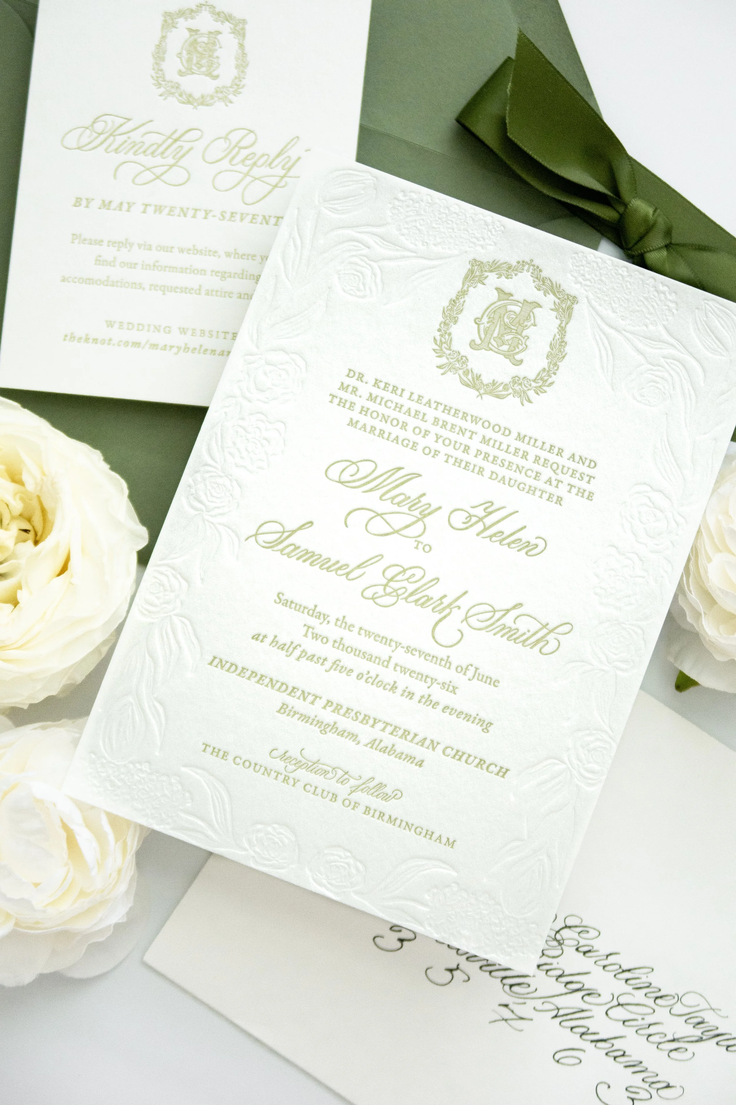 Elegant wedding invitation with embossed floral border and gold lettering, accompanied by cream-colored roses and a green ribbon.