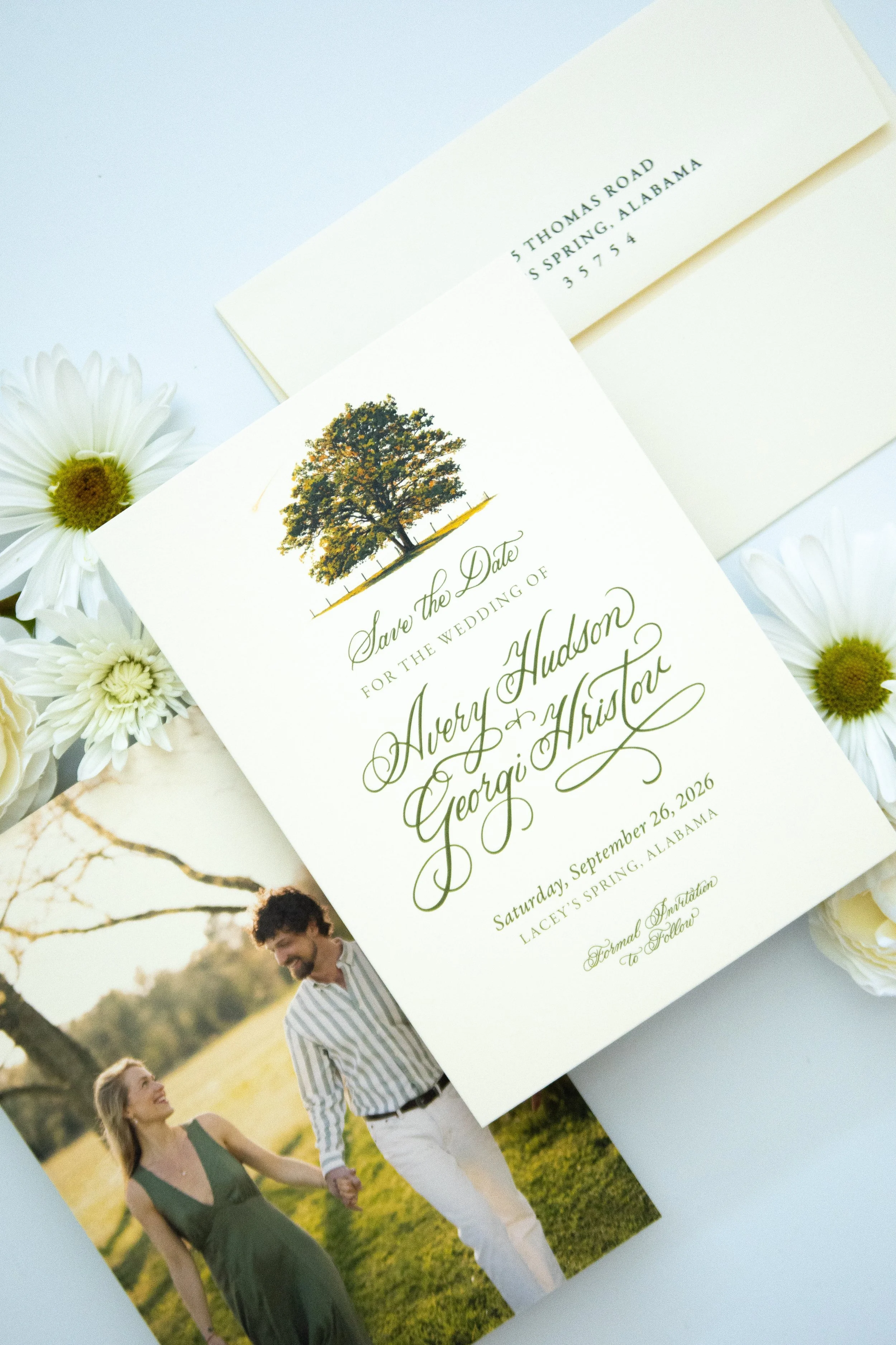 Wedding invitation card with a tree illustration and elegant green script text, surrounded by white daisies. A couple is walking hand in hand outdoors on a grassy field, with sunlight and trees in the background.