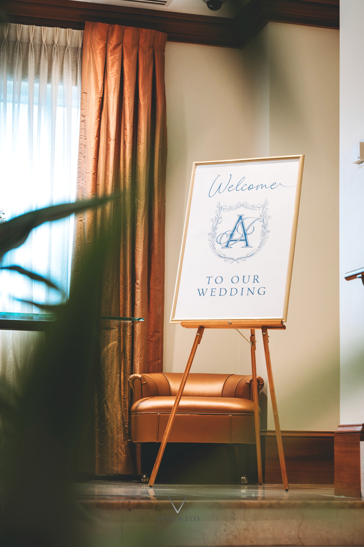 A welcome sign on an easel reads 'Welcome to our wedding' with initials 'J & A' in a decorative frame. The setting has warm-toned curtains, a brown armchair, and soft lighting.