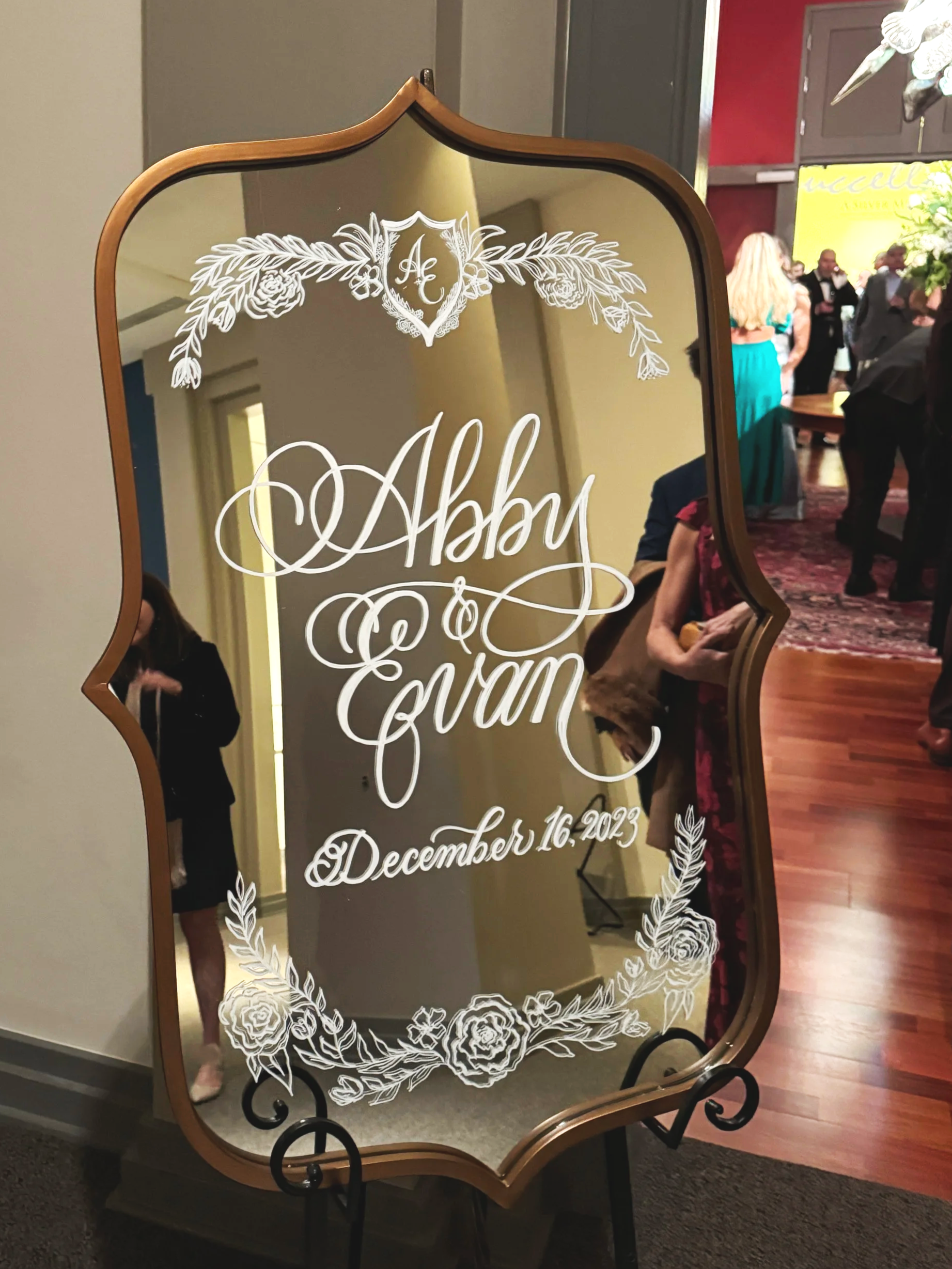 Decorative mirror with white floral design and text reading 'Aubrey & Evan, December 16, 2023,' at an indoor event with people in the background.