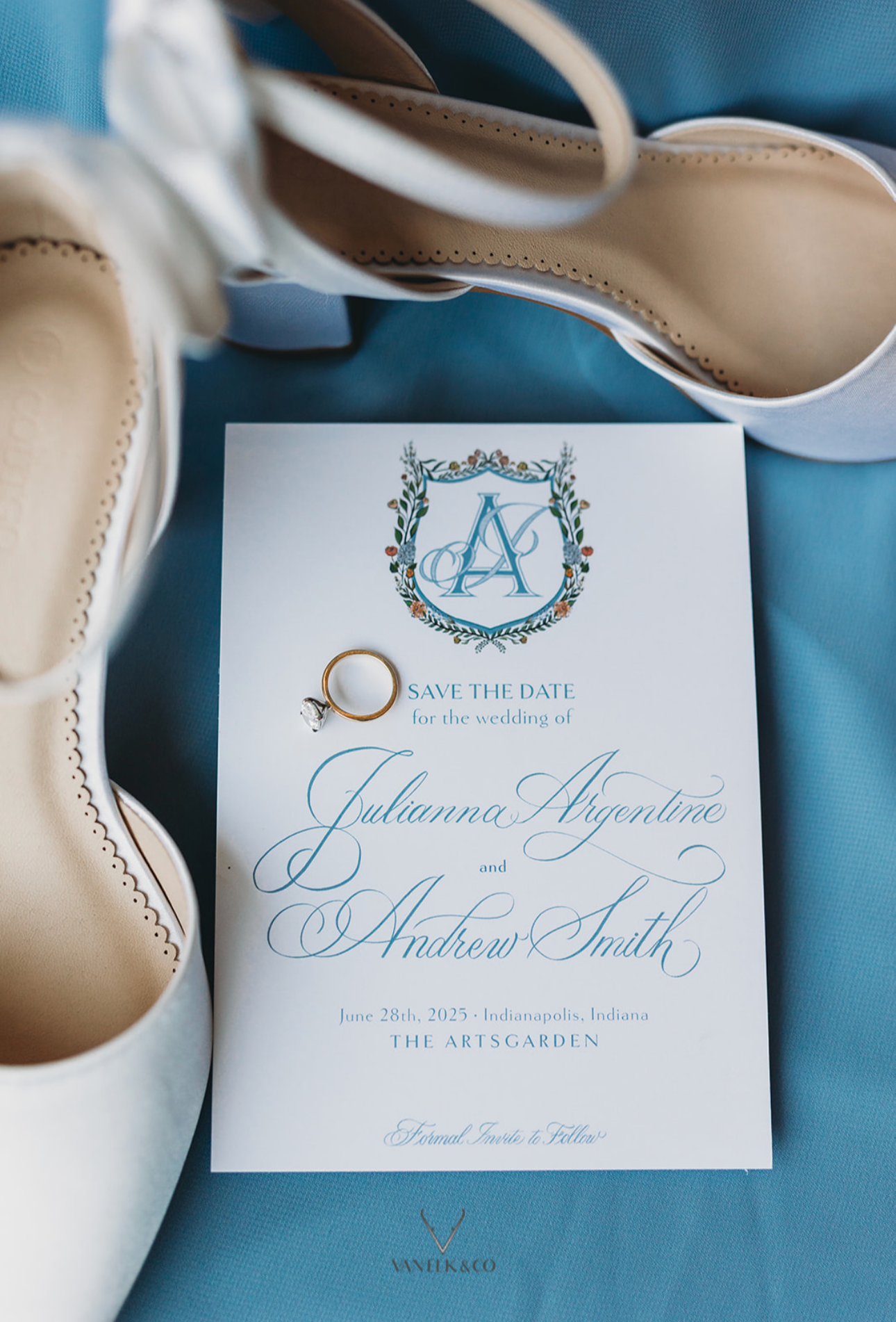Wedding save-the-date card with elegant script, featuring wedding details and a gold ring, surrounded by a pair of ivory shoes on a blue surface.