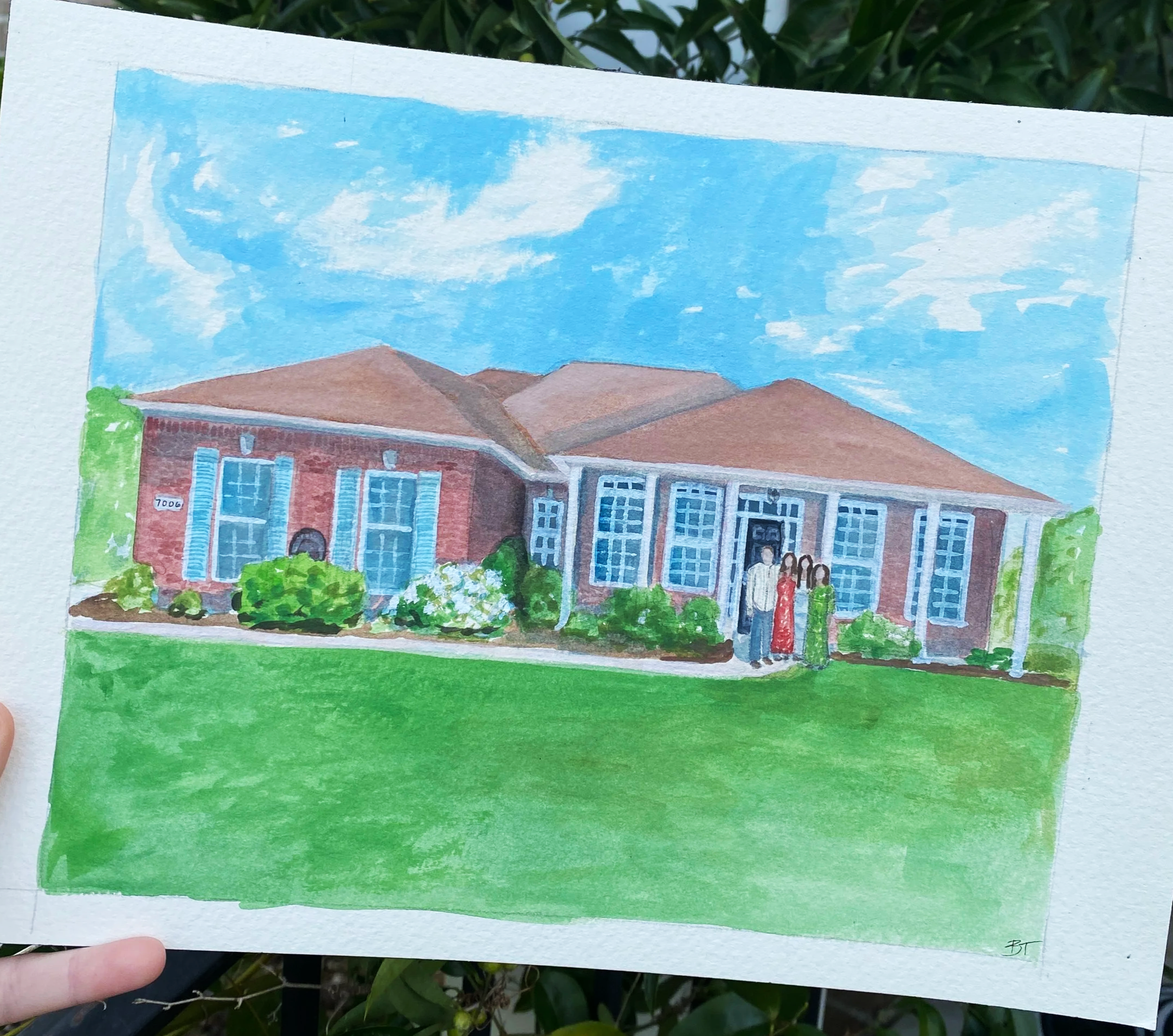 Hand holding a watercolor painting of a building with red brick walls, blue windows, and a sloped roof, set against a sky with clouds and greenery at the bottom.