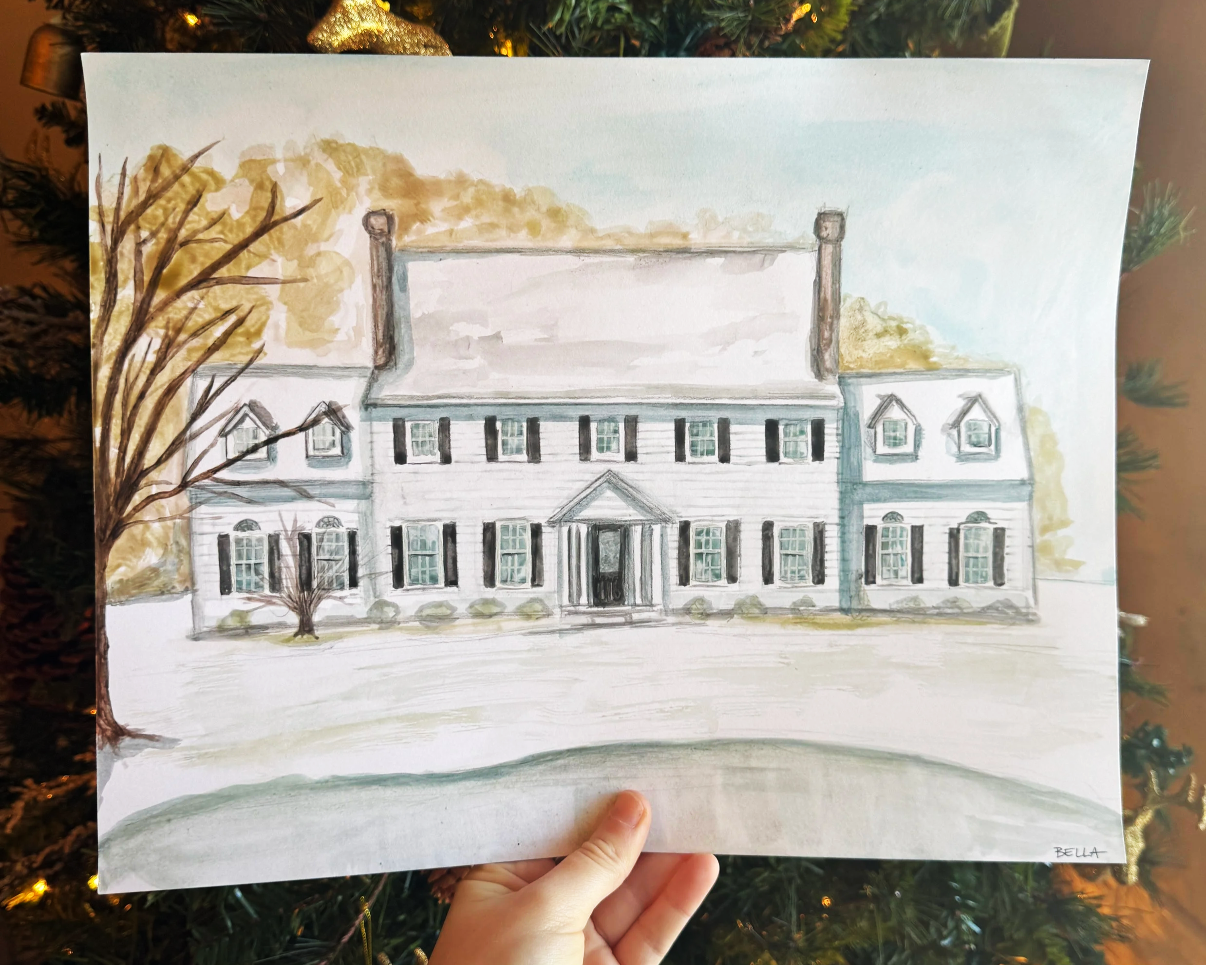 A watercolor painting of a large house with multiple windows, a porch, and a steep roof, surrounded by trees with autumn-colored leaves, depicted on a piece of white paper held by a person's hand, with a Christmas tree in the background.