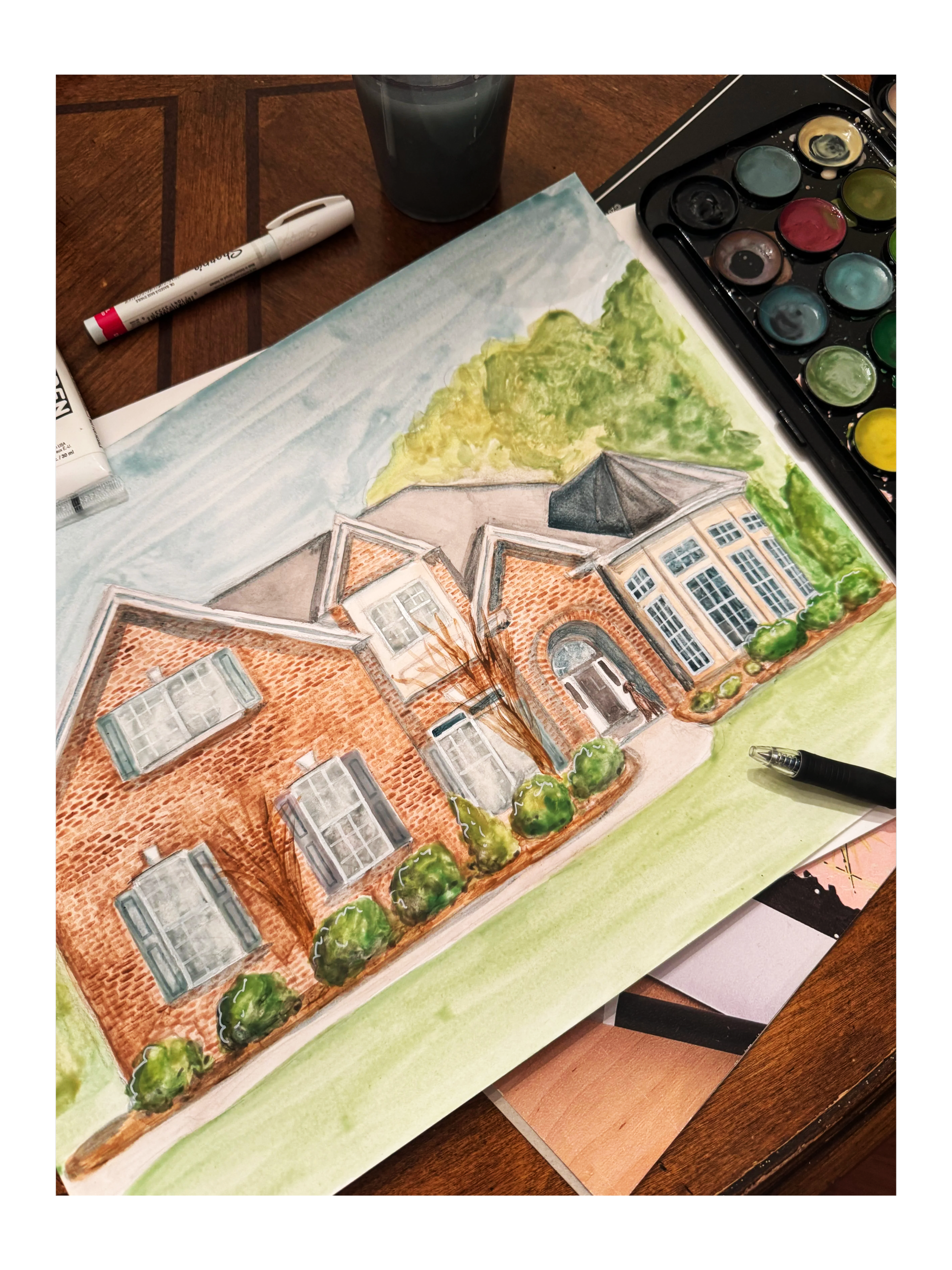 A watercolor painting of a brick house with large windows, surrounded by trees and grass, with art supplies and watercolors nearby.