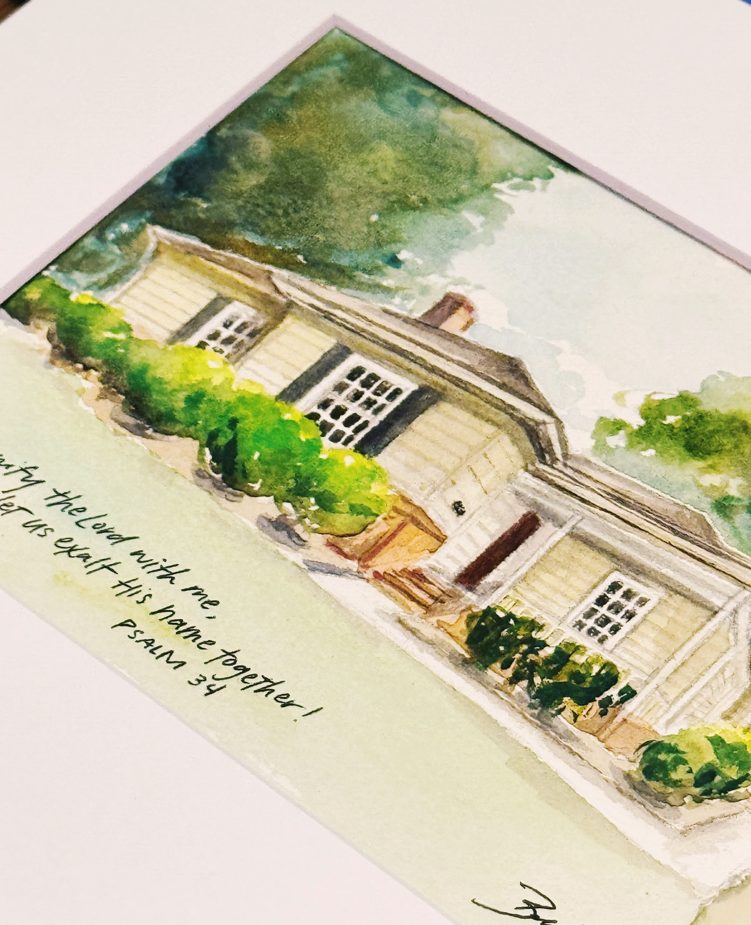 Watercolor painting of a house with light yellow walls, white-framed windows, and a dark roof, surrounded by green foliage and trees, with part of a handwritten note visible at the bottom.