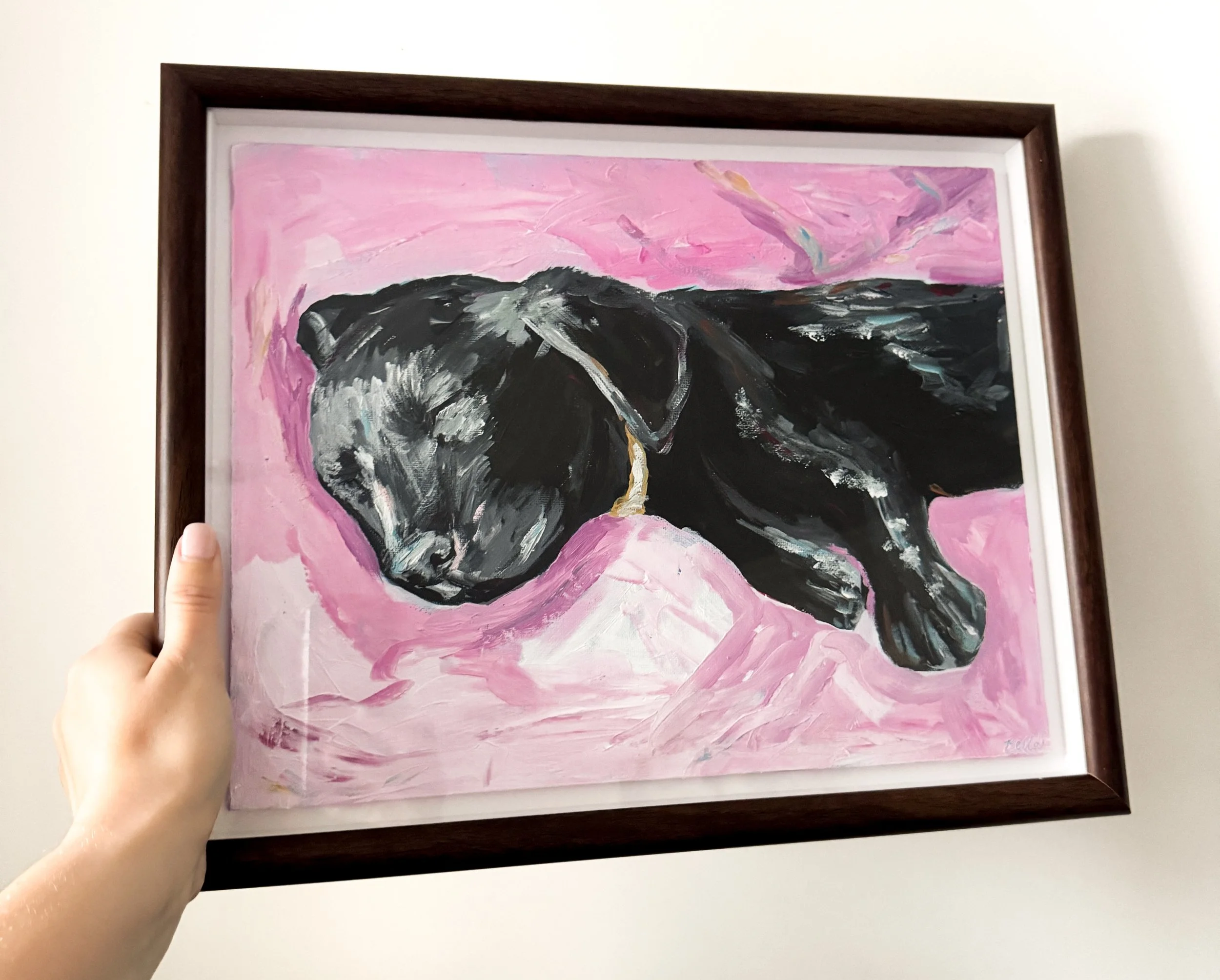 A person holding a framed abstract painting of a black sleepy puppy against a pink background.