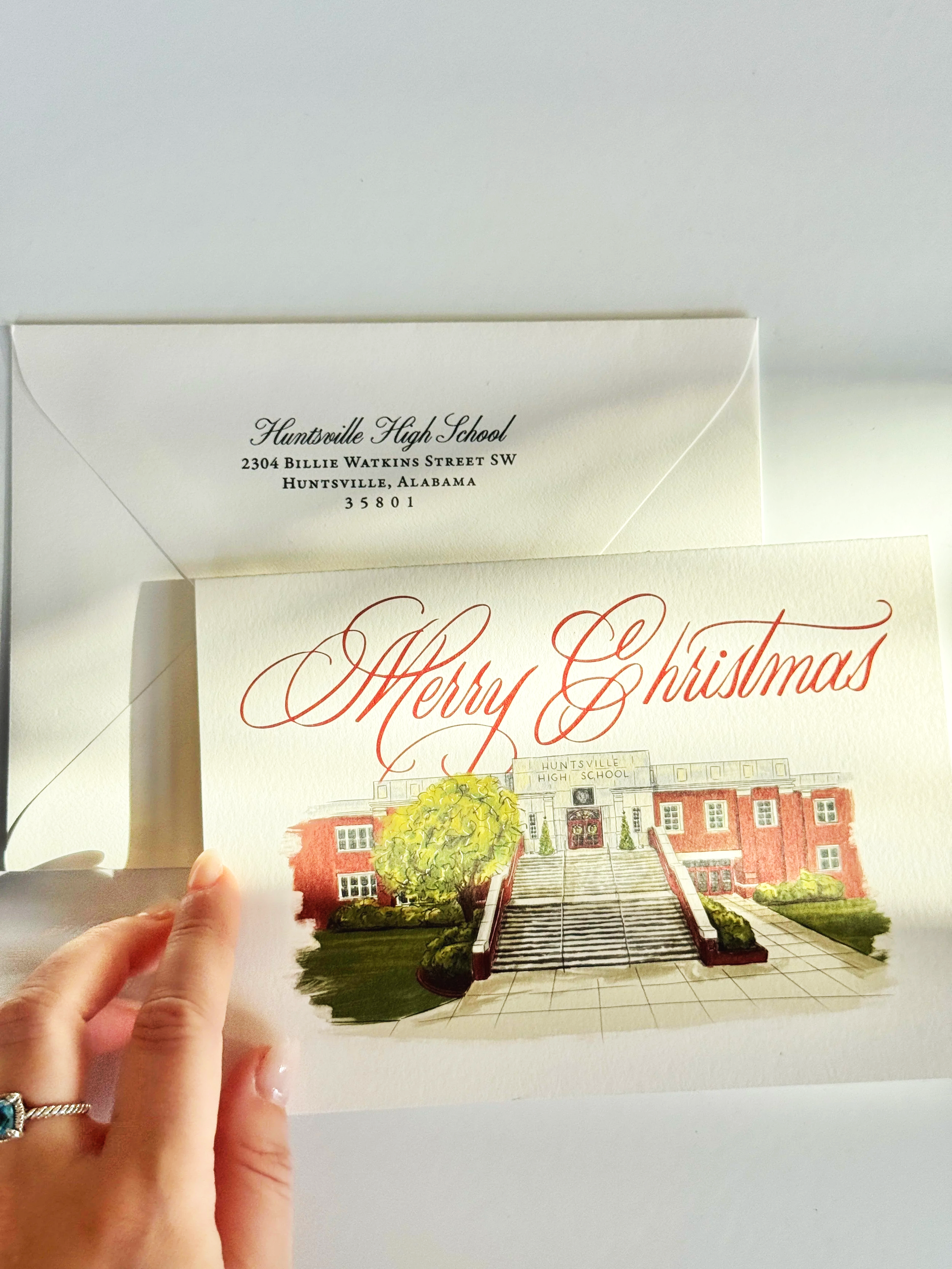 A person holding a holiday greeting card featuring an illustration of Huntsville High School, with the words "Merry Christmas" written in red in a script font.