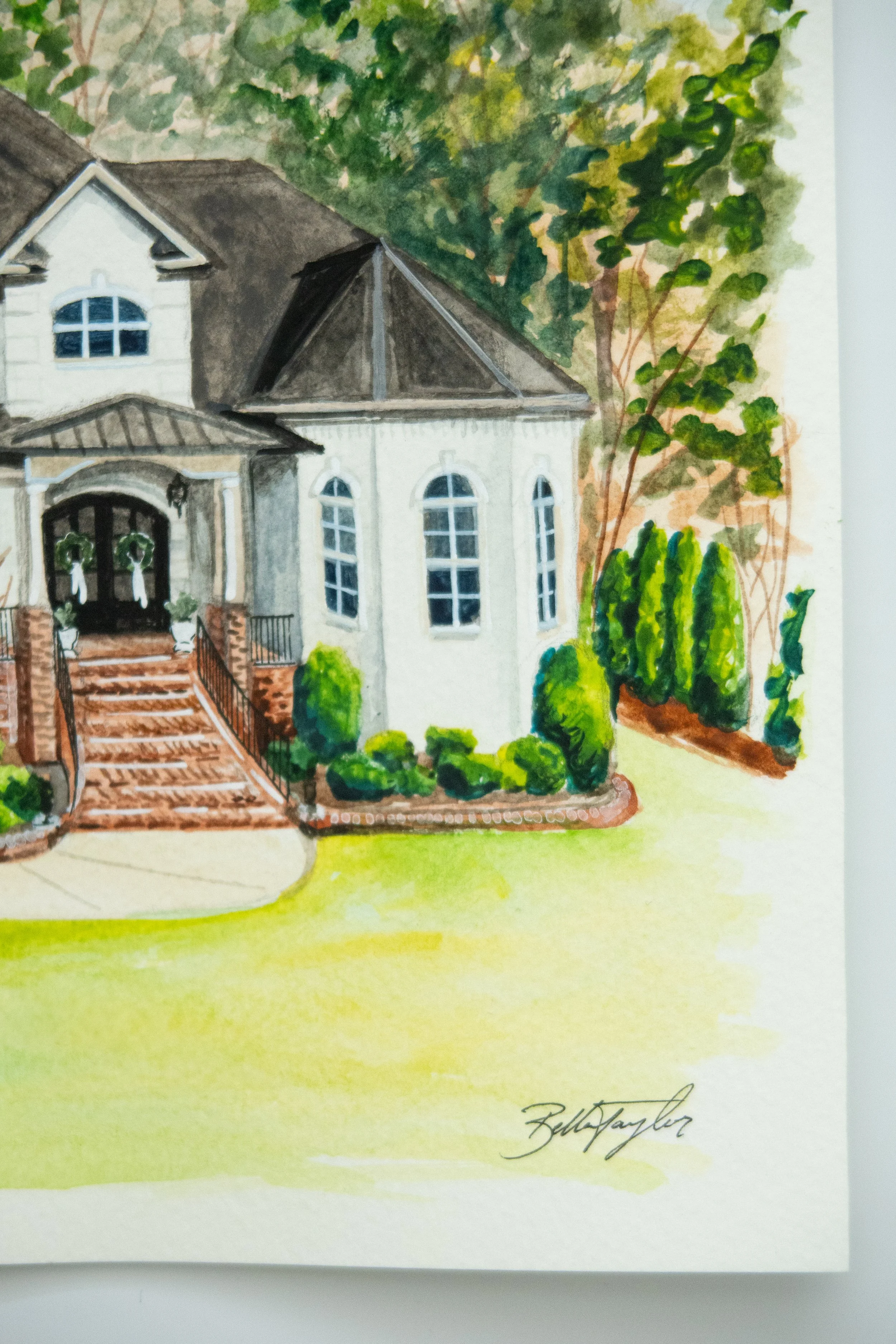 Watercolor painting of a house with brick steps leading to a front porch decorated with garland, surrounded by green shrubs and trees, signed by Bella Taylor.