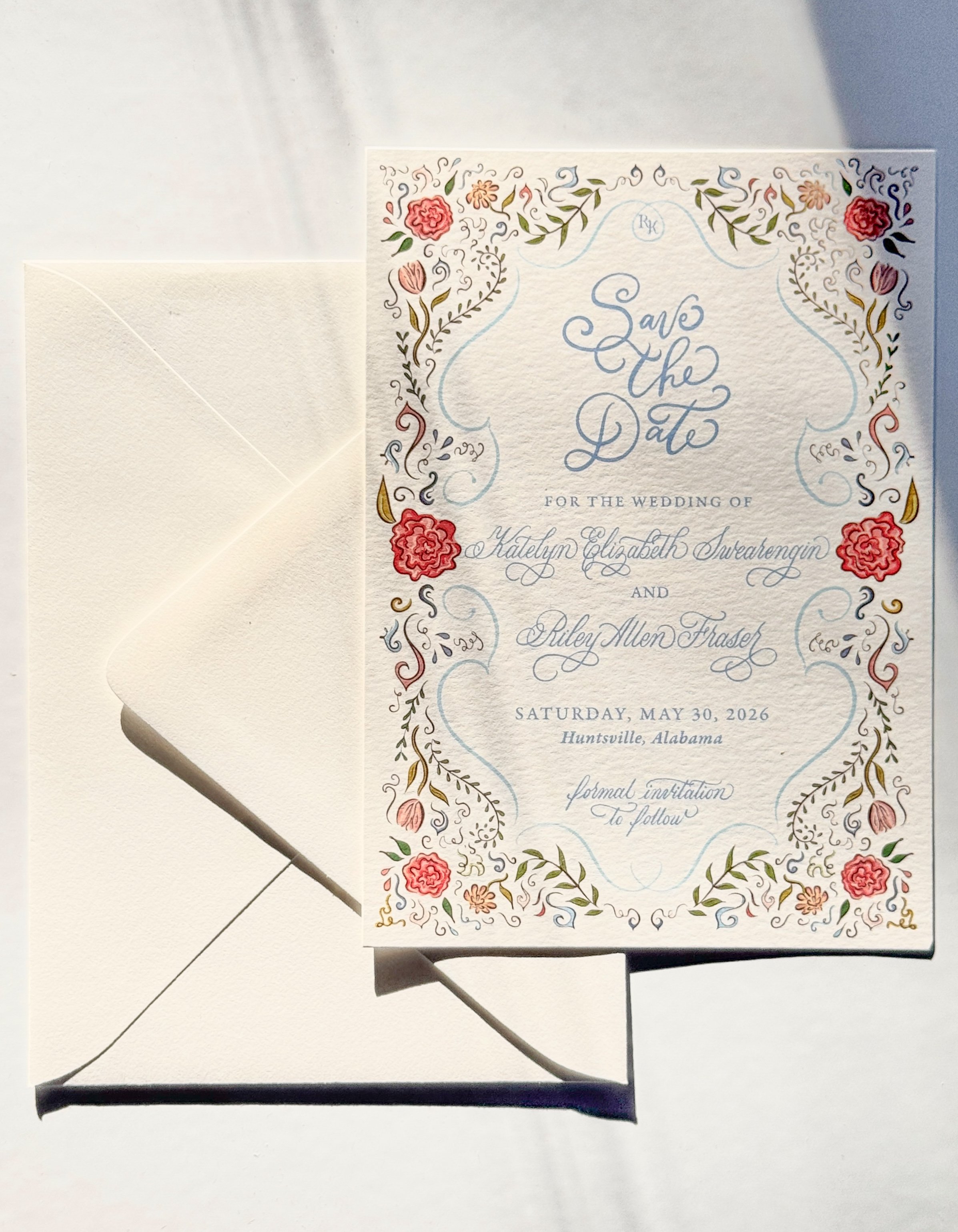 A floral wedding invitation with decorative border details and elegant blue script text, placed on an off-white background with an envelope partially visible.