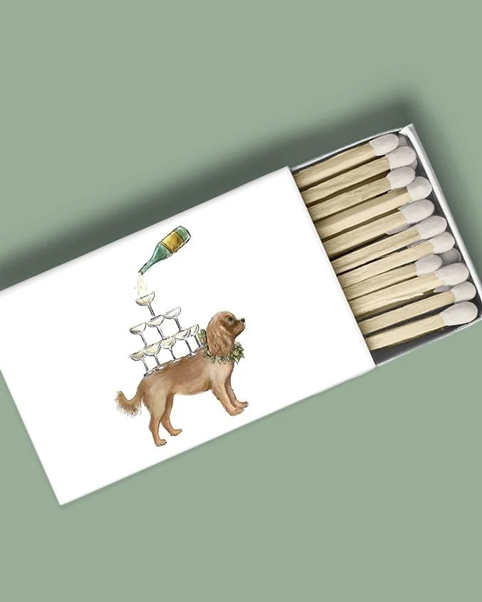 Box of matches with a drawing of a dog on the lid, featuring a champagne bottle pouring into a glass on its back, and four champagne glasses stacked in a pyramid on its body, with a green collar or wreath around its neck.