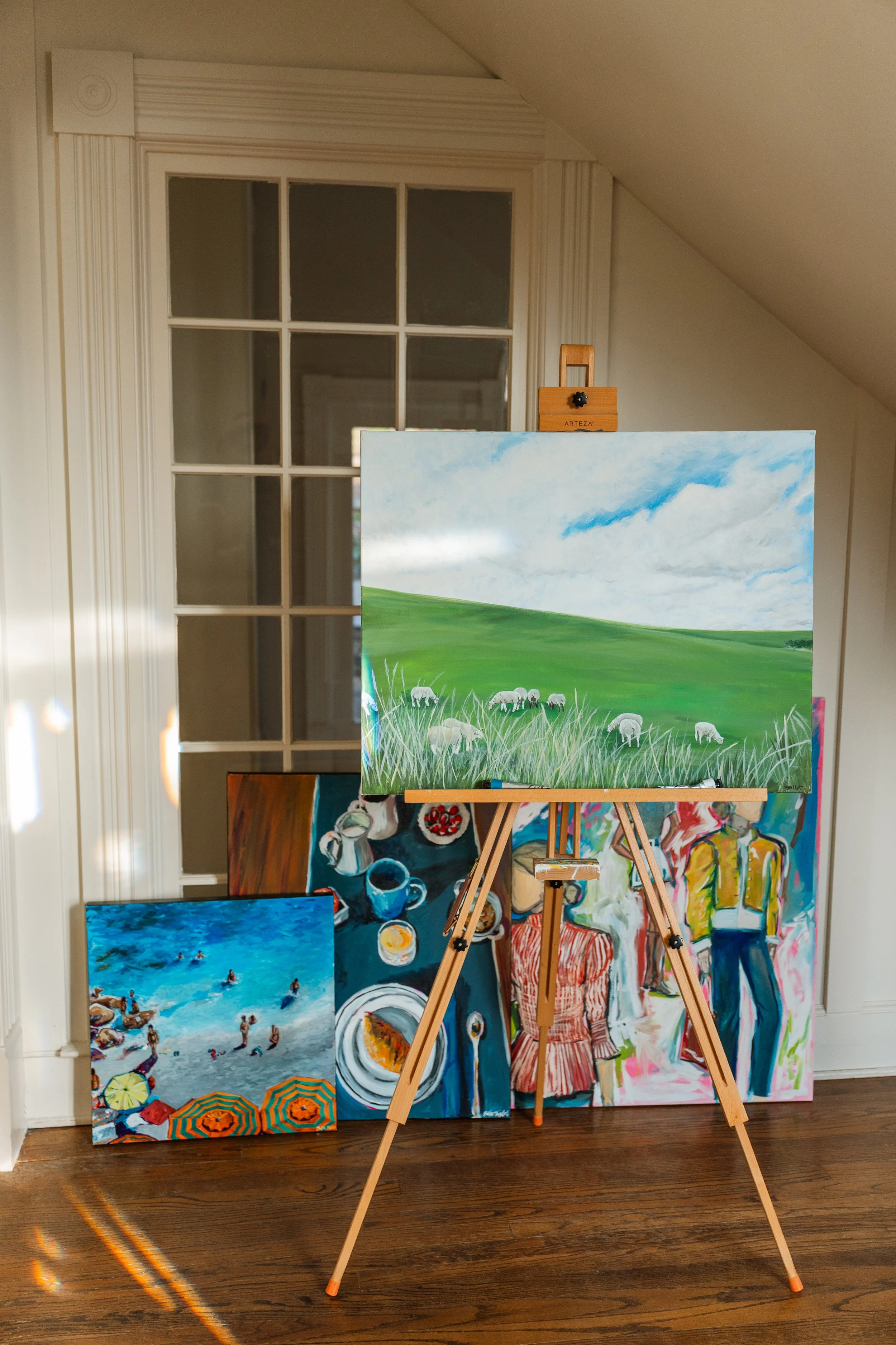 An art studio with a wooden easel holding a landscape painting of green hills and a blue sky with clouds. Several other colorful paintings are leaning against the wall, including a beach scene, a still life of cups and plates, and a figure painting. 