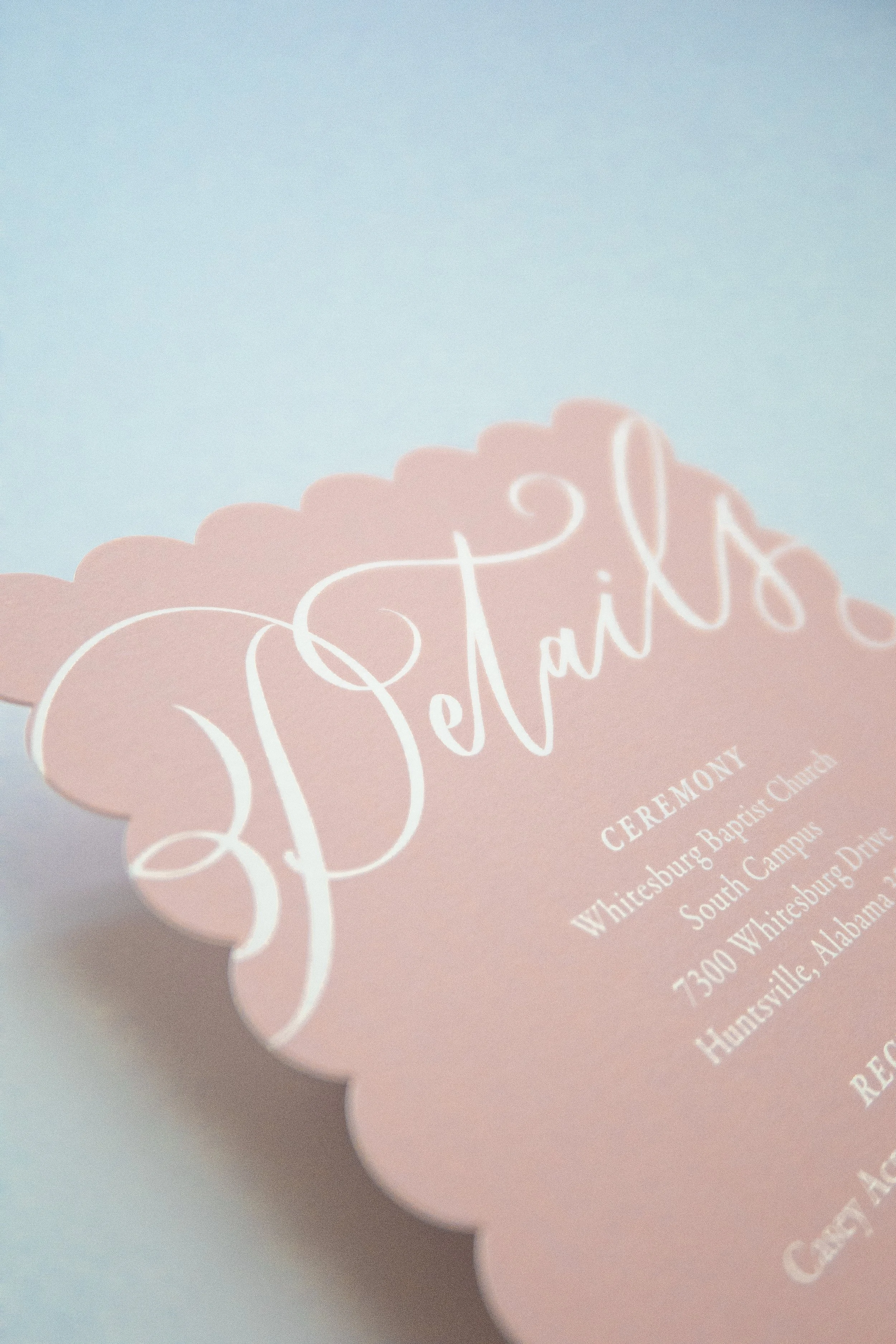 Close-up image of a pink scalloped design featuring decorative white script text for a wedding ceremony card with details about the location and date.