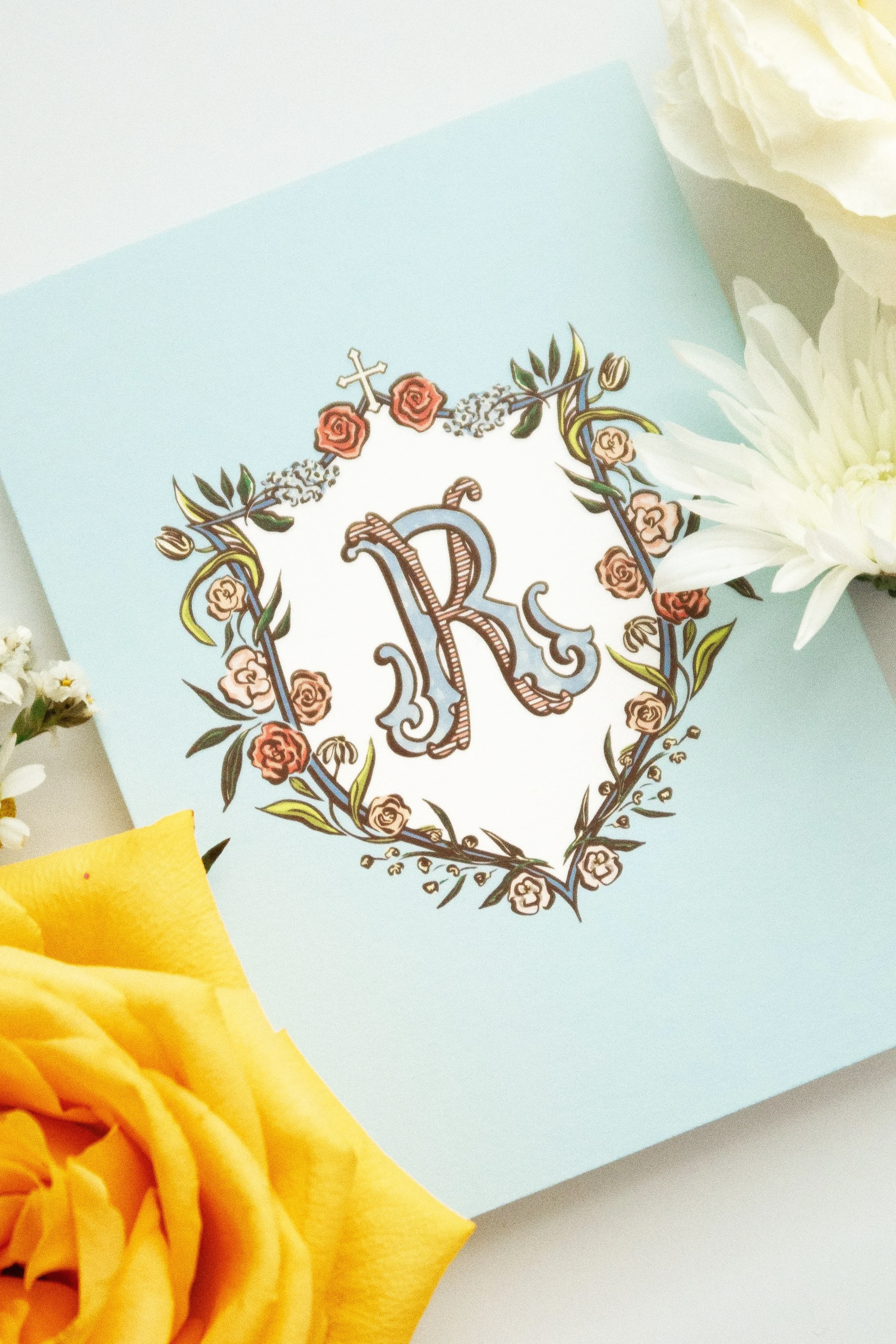 Close-up of a decorative paper item with floral and letter design, surrounded by yellow and white flowers on a pastel blue background.