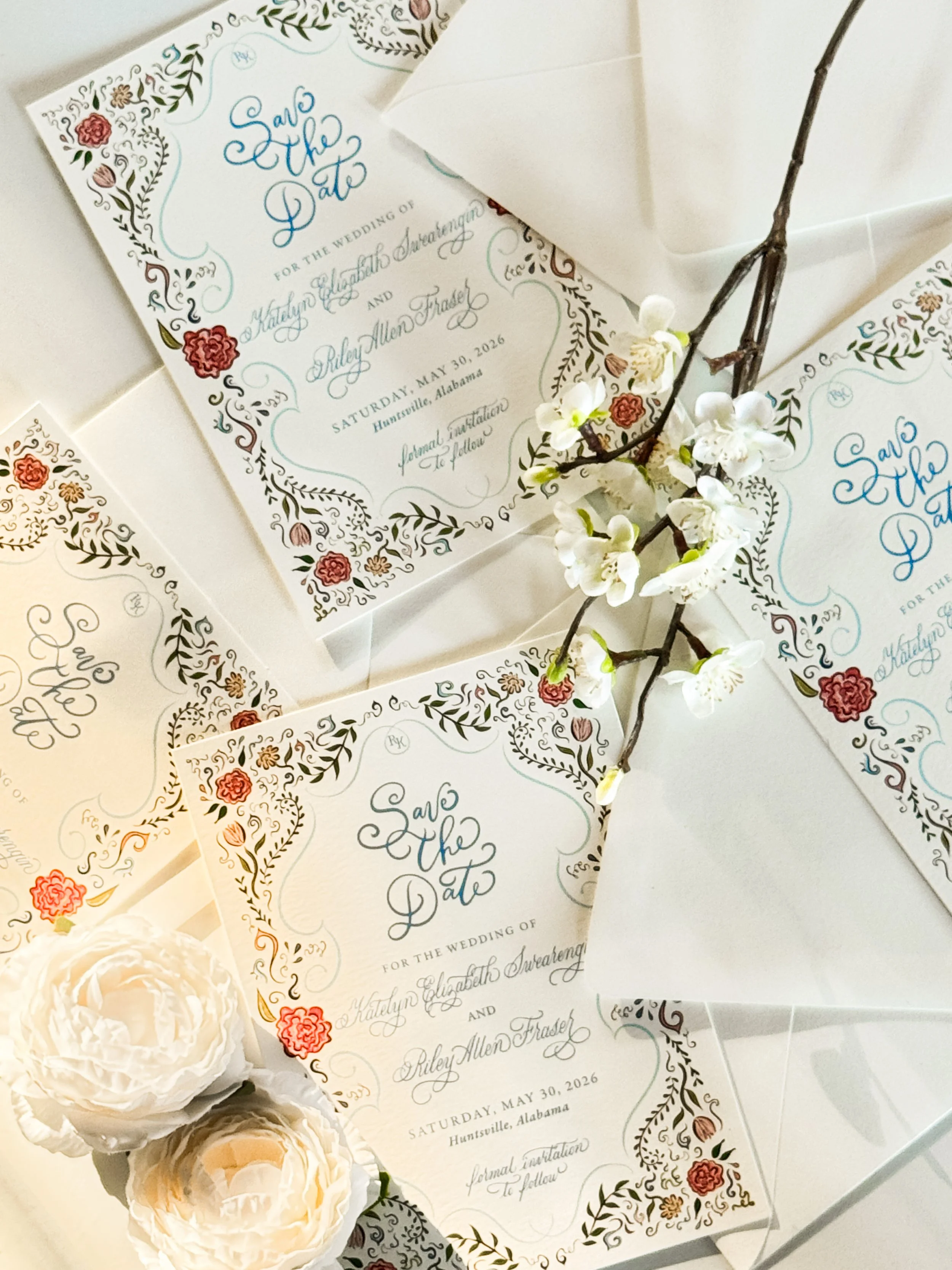 Multiple wedding invitations with floral designs and the words "Save the Date" in elegant script, accompanied by white and cream roses and a branch of white blossoms.