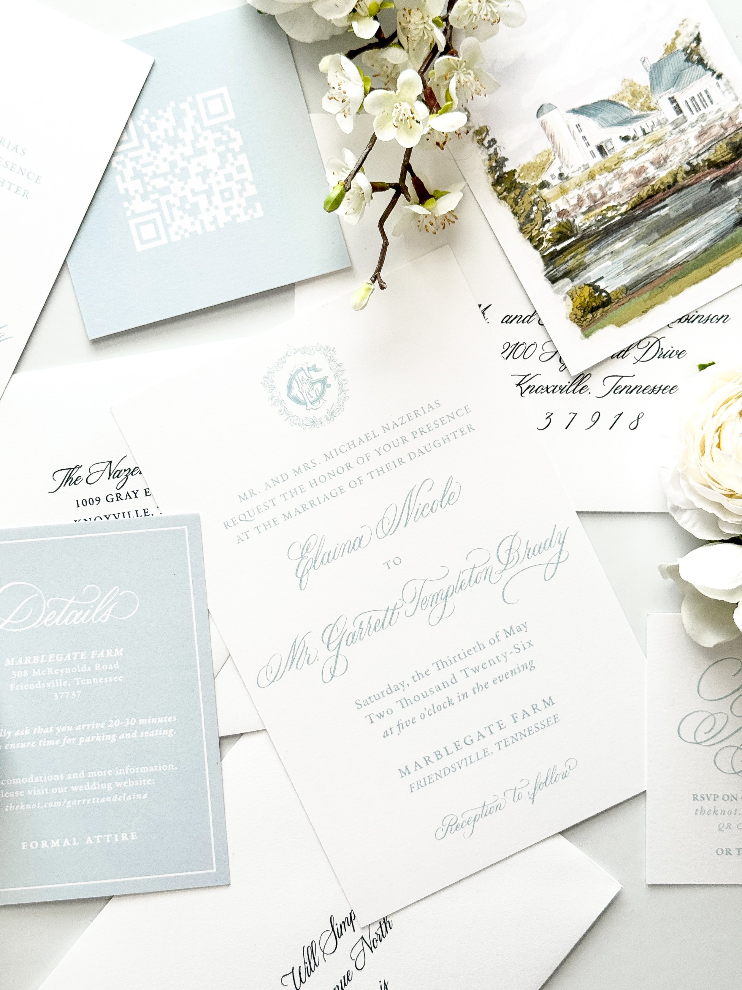 Multiple wedding invitation cards with elegant calligraphy, one featuring a watercolor painting of a house near a river, accompanied by a sprig of white blossoms resting on top.