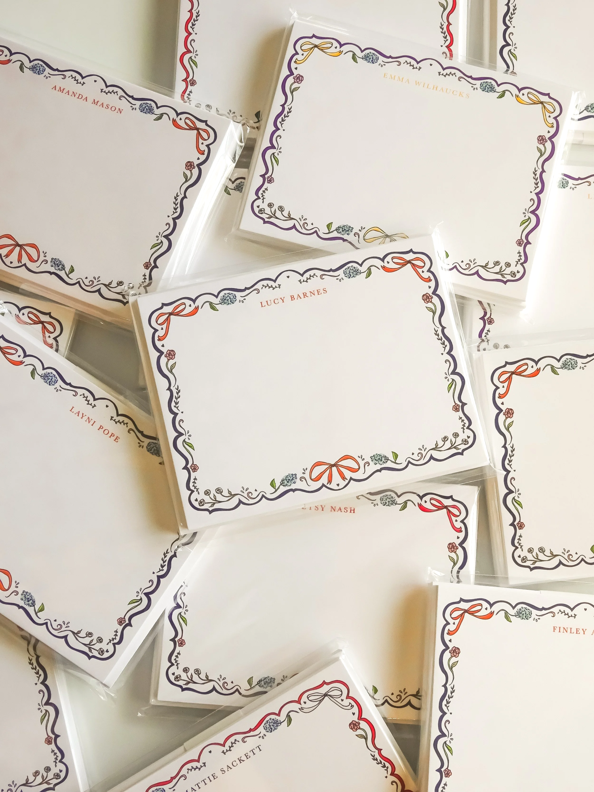Stacked custom stationery sets with white backgrounds, decorated with floral and bow border designs, featuring names.