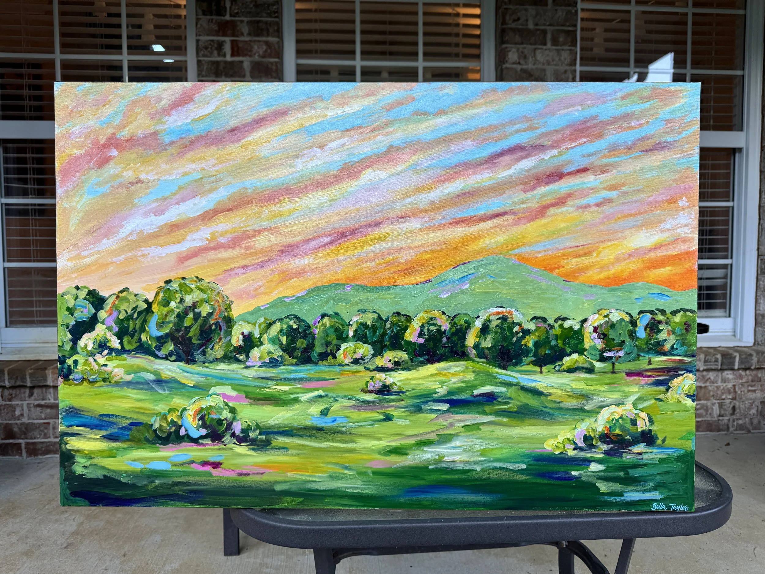A colorful landscape painting depicts a vibrant sunset over green hills and trees, with a sky filled with shades of pink, orange, yellow, blue, and purple.