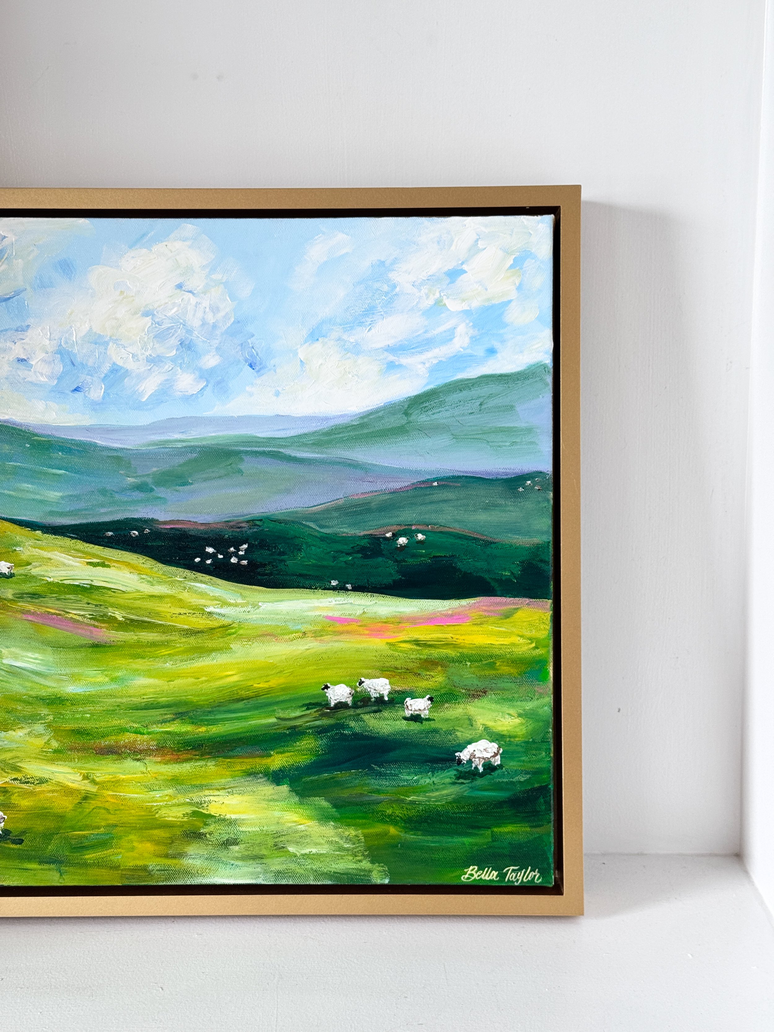 A landscape painting depicting green rolling hills, a blue sky with white clouds, and a few white sheep grazing in the foreground, signed by Bella Taylor.