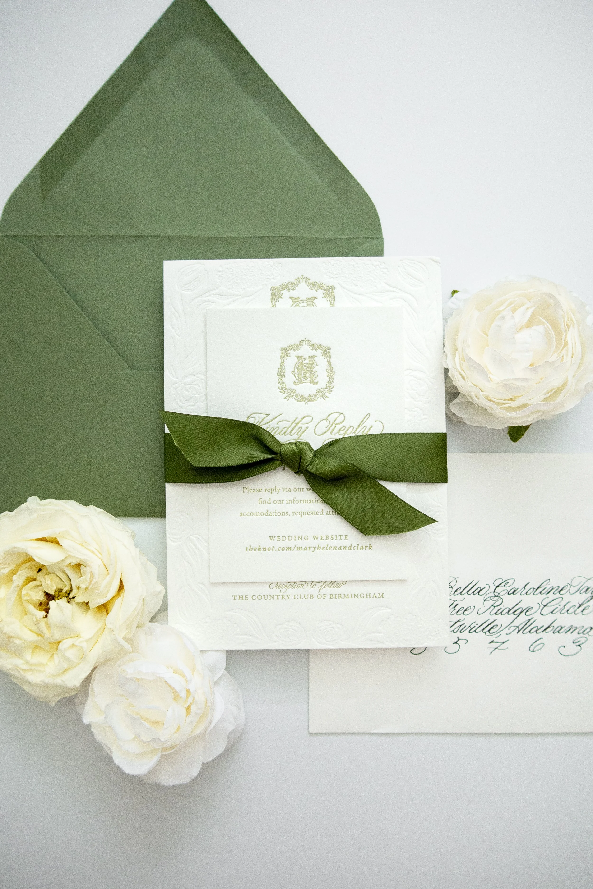 Wedding invitation card with cream-colored roses and a dark green ribbon, placed inside a dark green envelope. The card has gold embossed details and elegant script text.