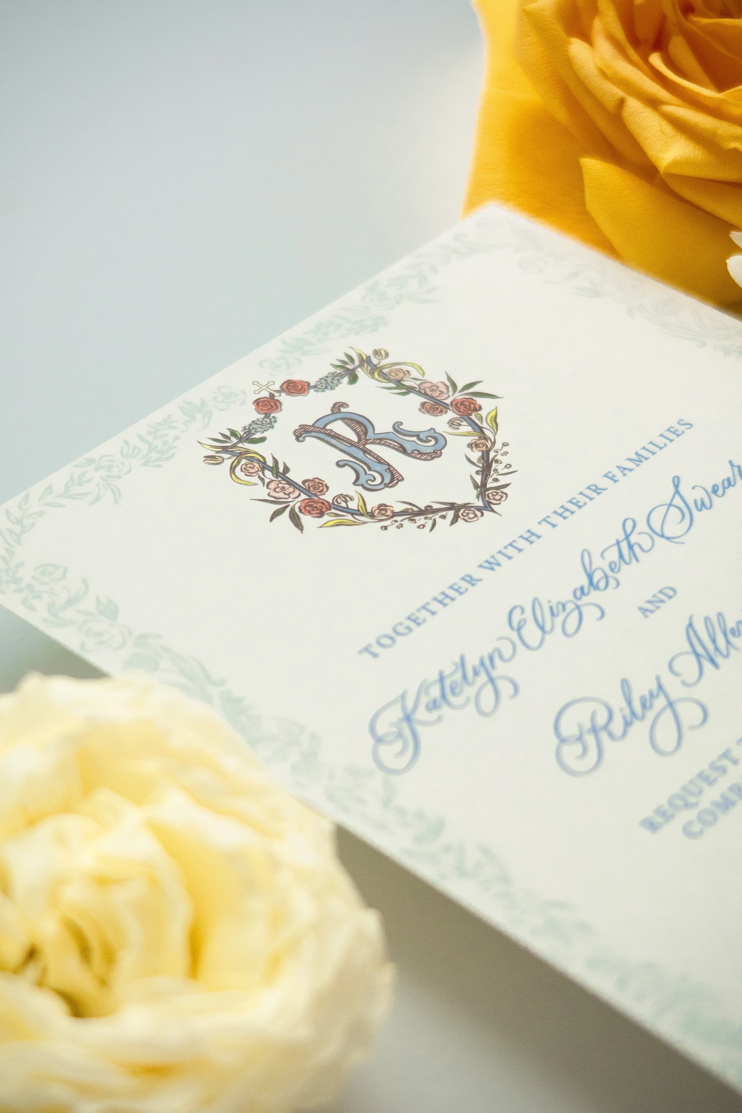 Close-up of a wedding or special occasion invitation or card with a decorative crest or emblem, floral border, and cursive text, placed on a surface next to a cream-colored flower
