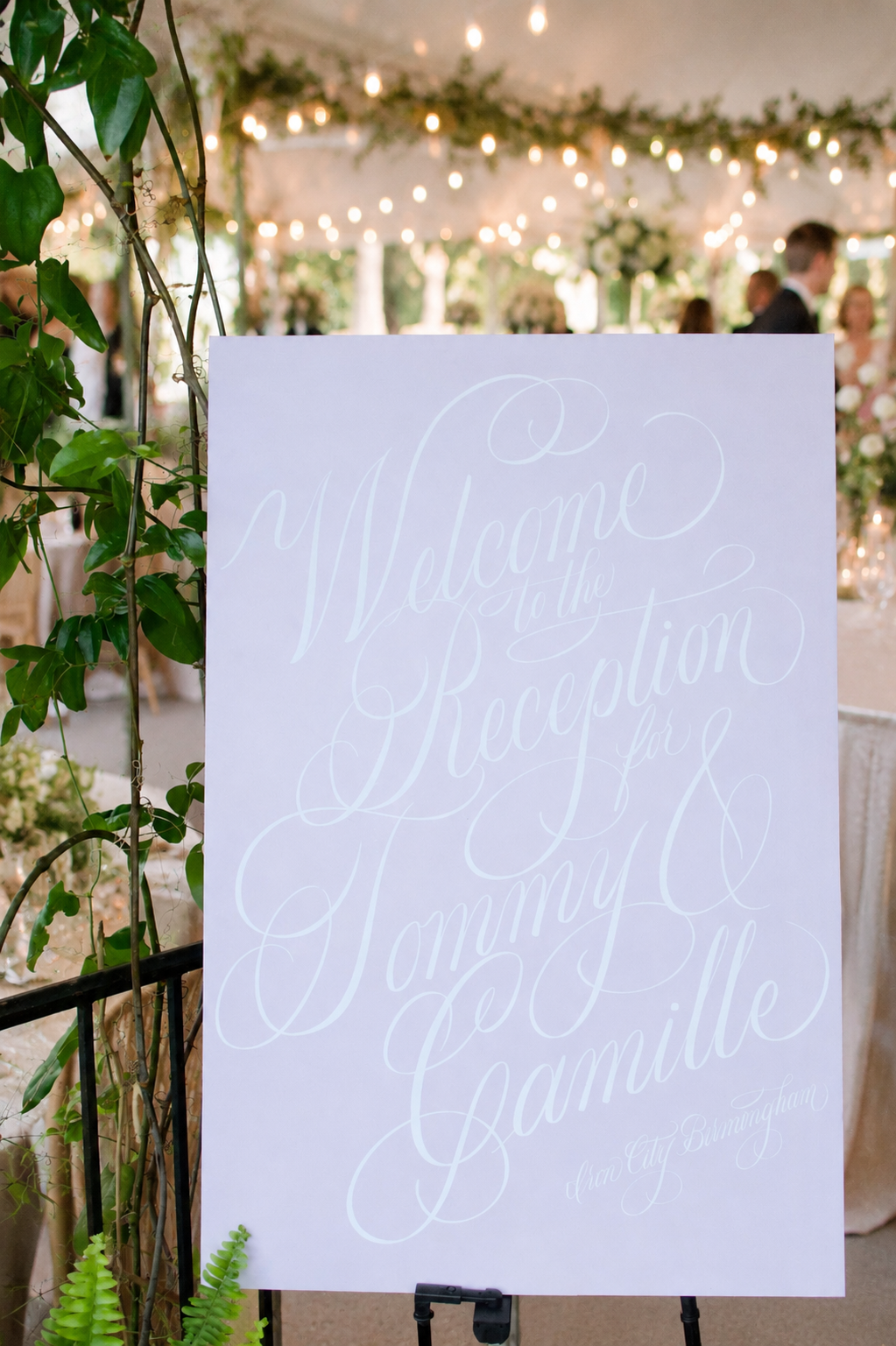 Wedding reception sign with elegant script reading 'Welcome to the Reception for Tommy & Camille, in Birmingham' indoors illuminated by string lights, decorated with greenery and flowers.