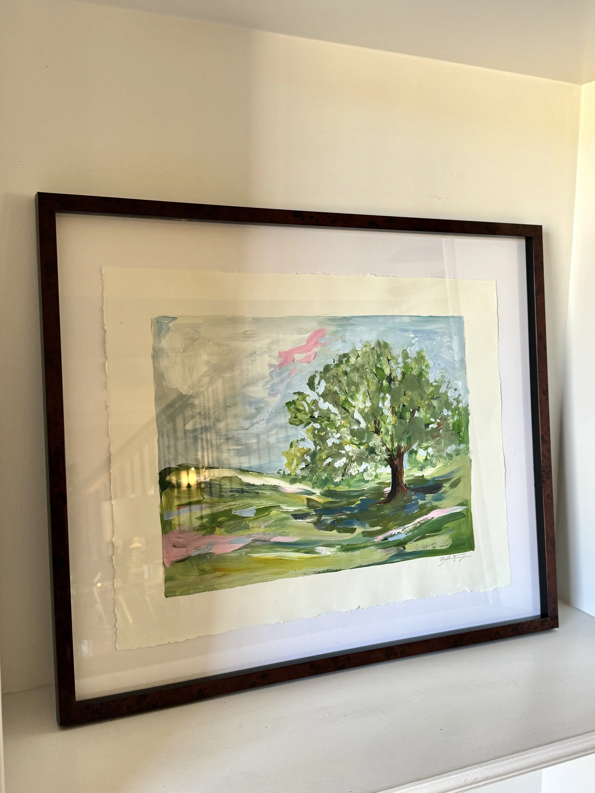 Framed watercolor painting of a tree with green leaves, pink flowers or blossoms, set against a blue sky with light clouds.