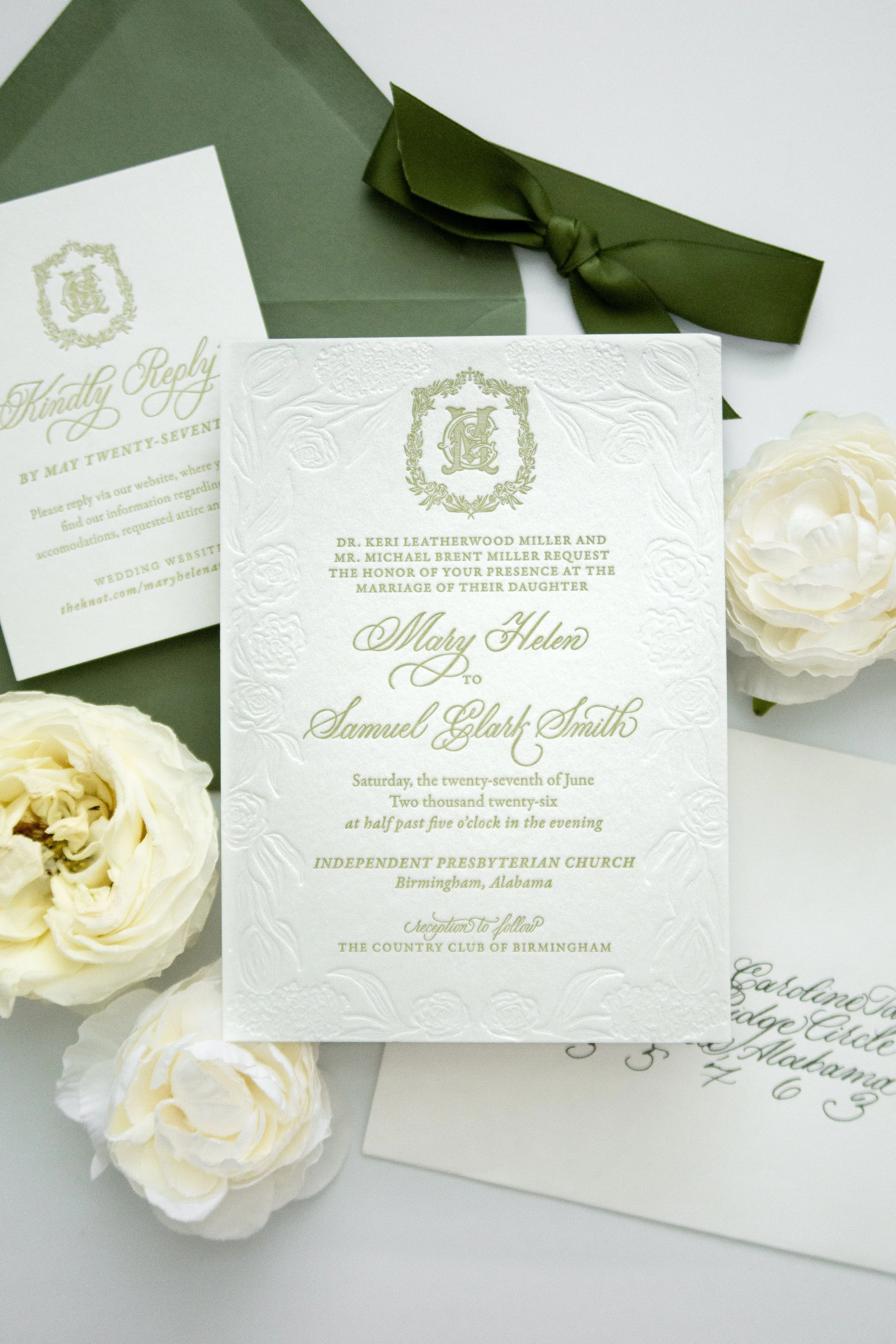 Wedding invitation card with embossed floral pattern and gold lettering, surrounded by white roses and placed on a white surface.
