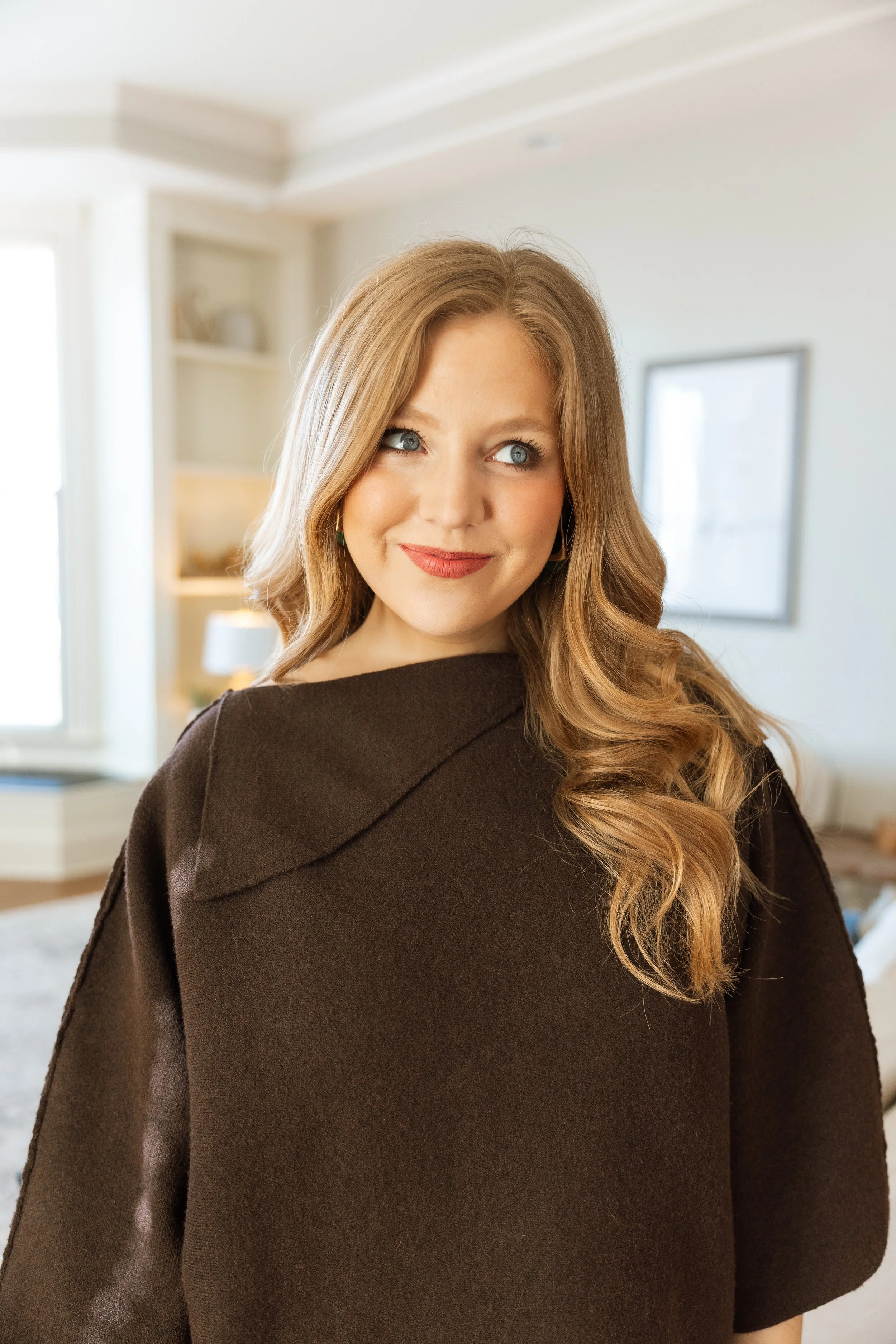 A woman with long, wavy, blonde hair and blue eyes, wearing a dark brown shawl and lipstick, standing inside a bright room.