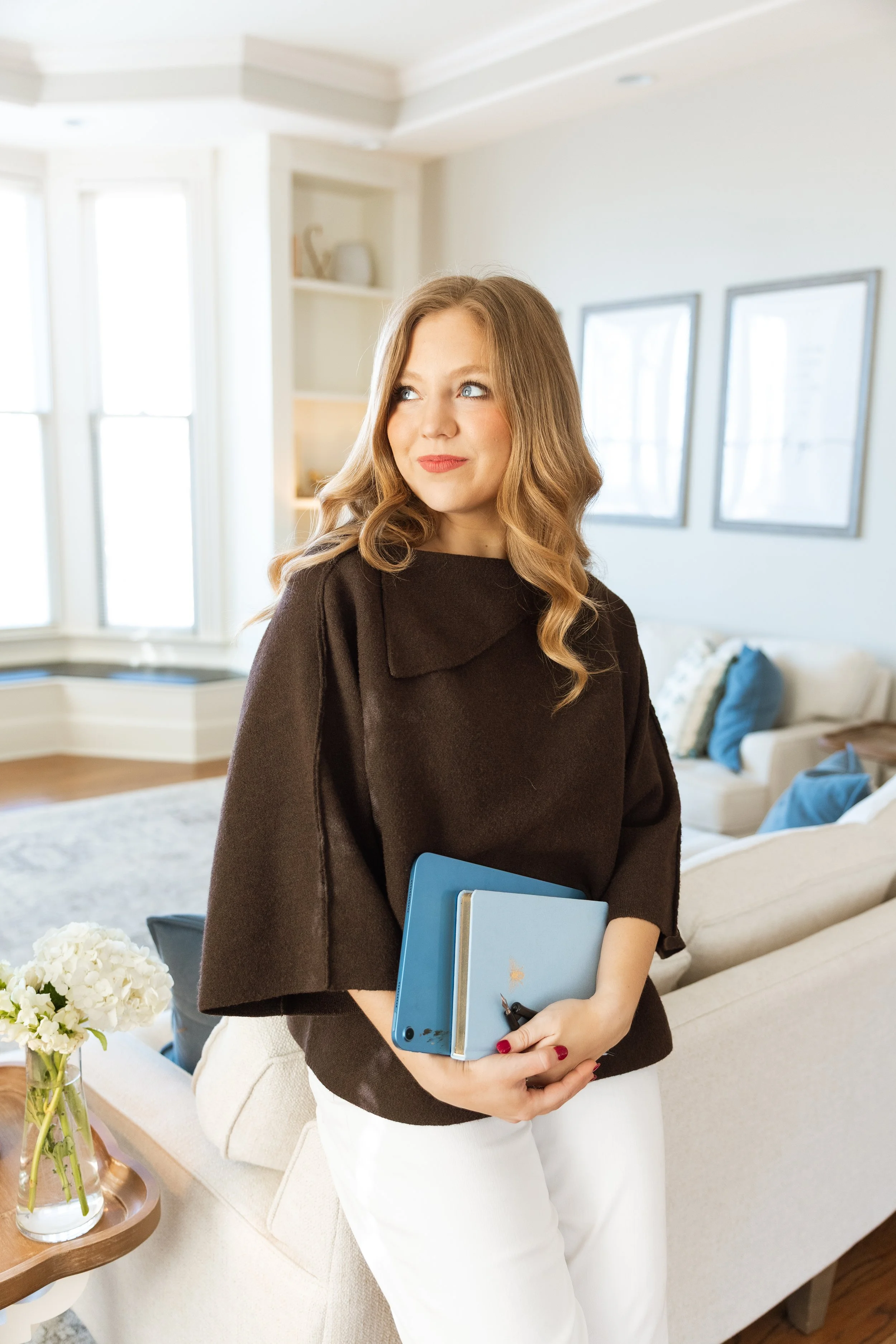 A woman with long wavy hair, wearing a dark brown top and white pants, holding a blue notebook and a smartphone, standing in a bright living room with large windows, a cream-colored sofa, and framed artwork on the wall.