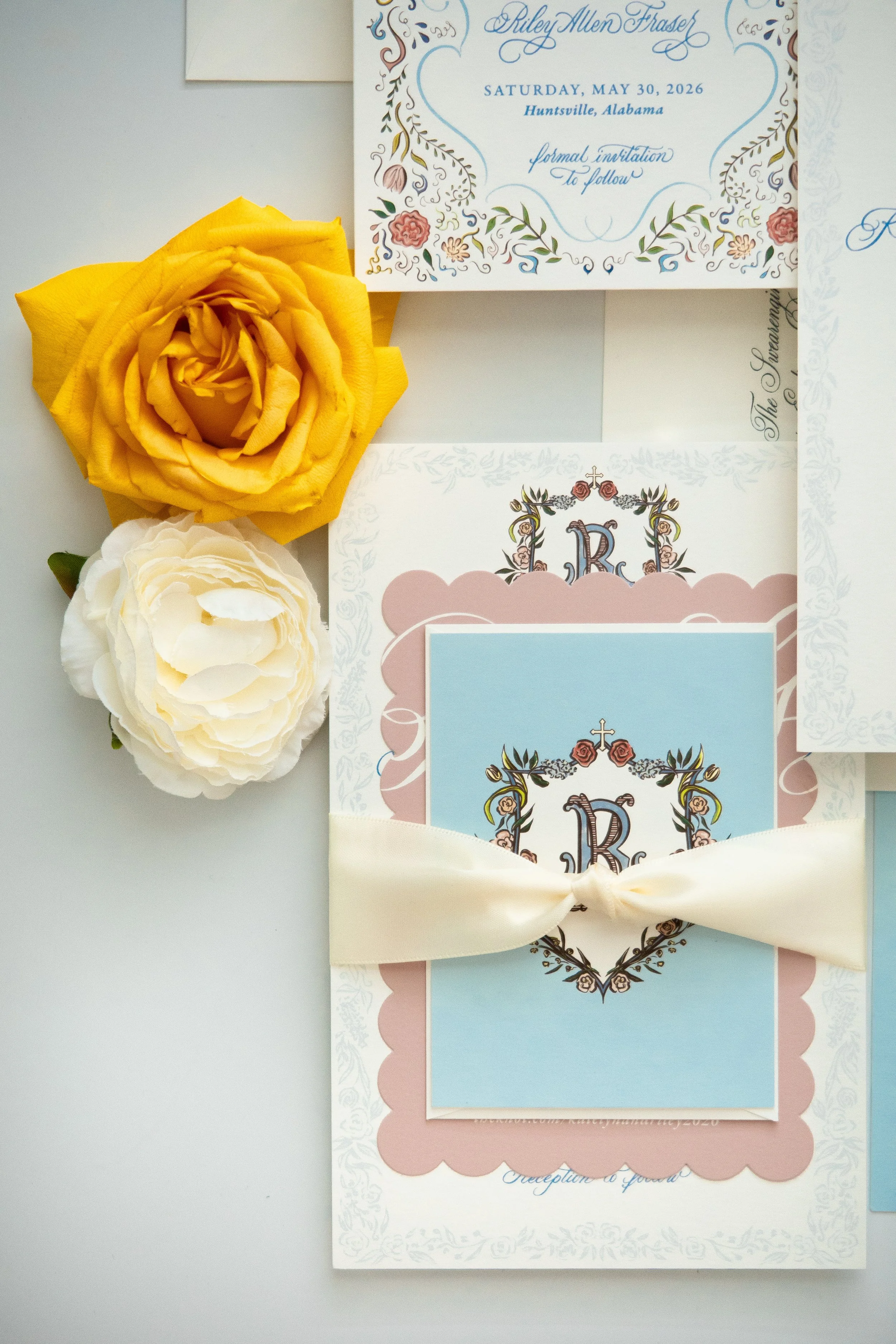 Wedding invitation card with floral and ornate design, featuring the initials 'R' and 'K', decorated with pink roses, purple flowers, green leaves, and a gold cross, tied with a cream ribbon. There are also two artificial flowers, a yellow rose and a