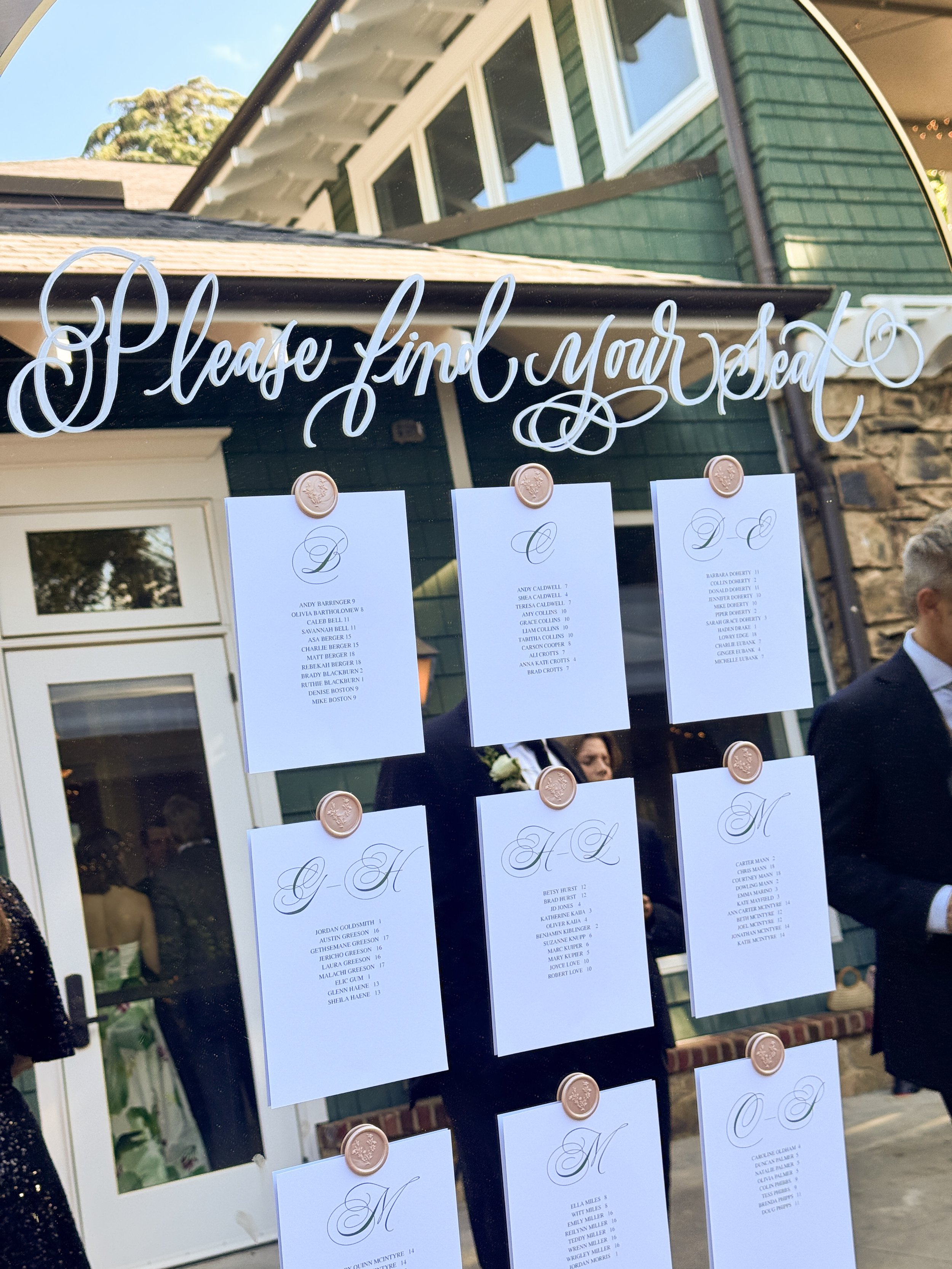 A mirror displays a seating chart with names and table numbers for a wedding or event, with the phrase 'Please find your seat' written in cursive at the top.