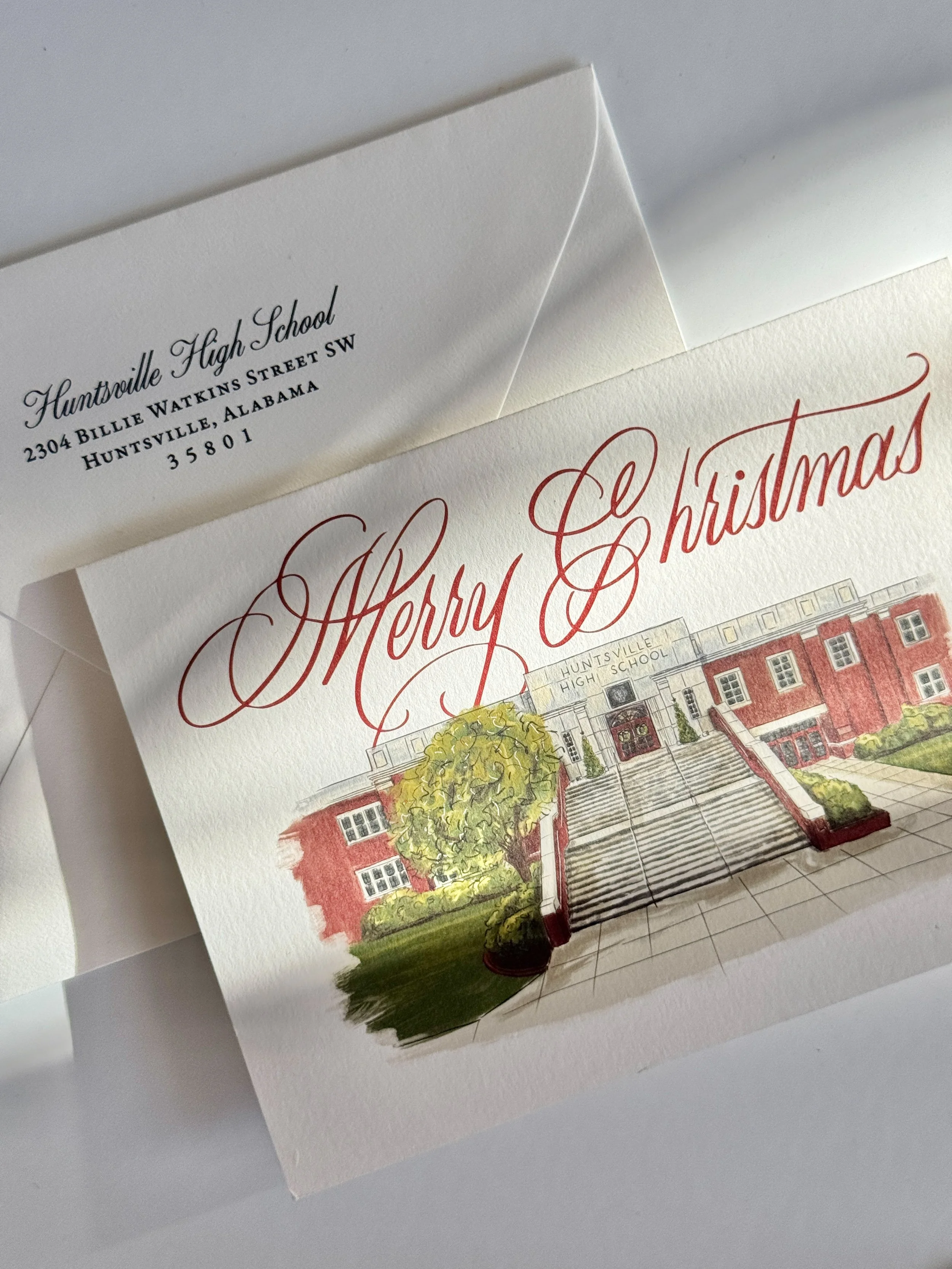 A Christmas card featuring an illustration of Huntsville High School, with red brick building, stairs, and trees in front, and the phrase 'Merry Christmas' in red cursive text.