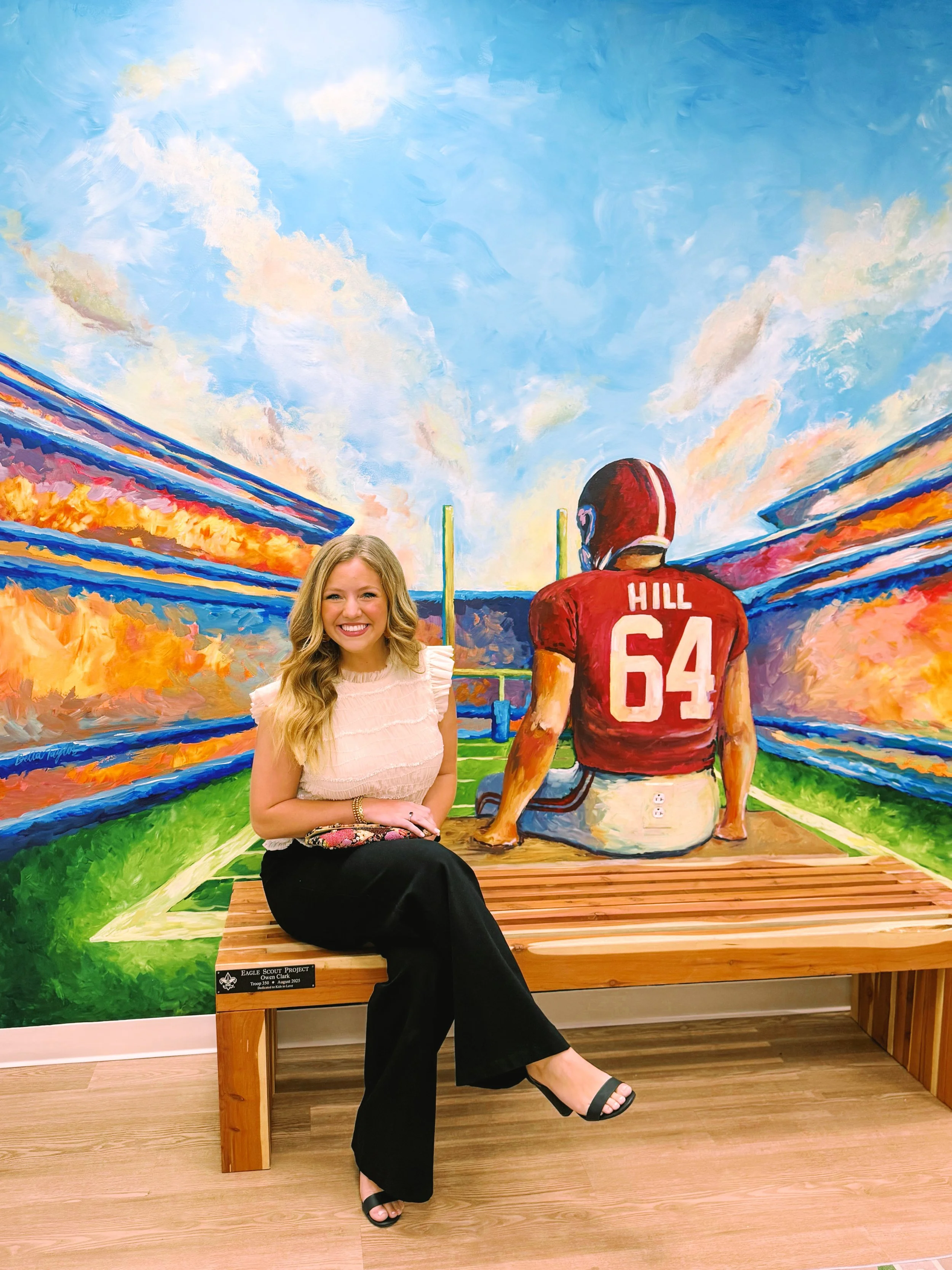 A woman with long blonde hair sitting on a wooden bench in front of a colorful mural of a football stadium scene with an athlete in a red jersey sitting on the ground with his back facing the camera. The woman is smiling and wearing a light pink top, black pants, and black sandals.